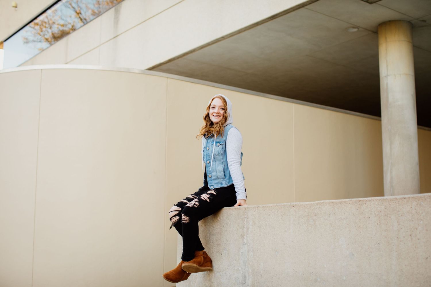 Young woman in denim vest and black ripped jeans sitting on a concrete wall smiling outdoors.