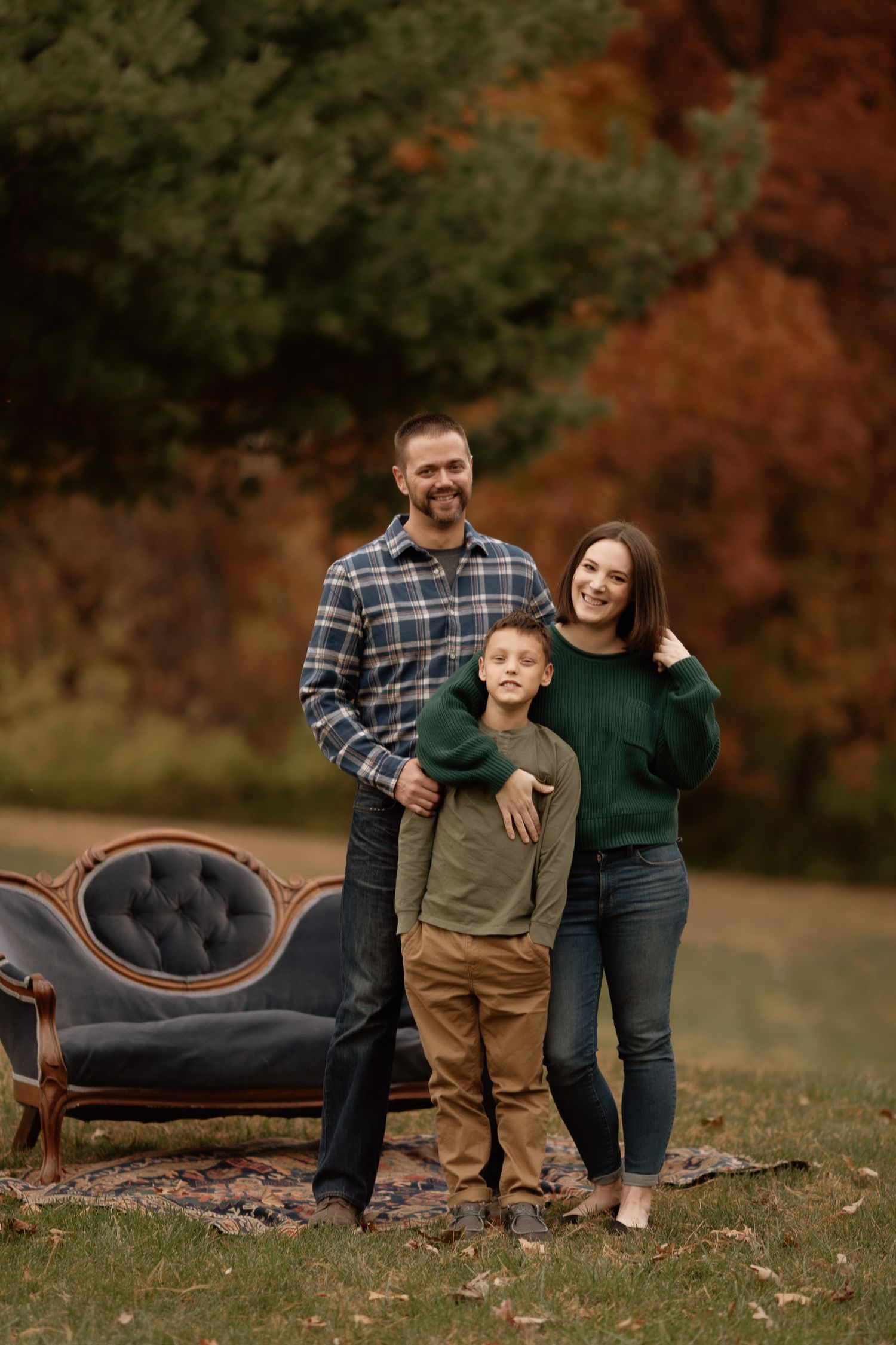 Family of three posing outdoors in autumn with a vintage tufted sofa behind them.