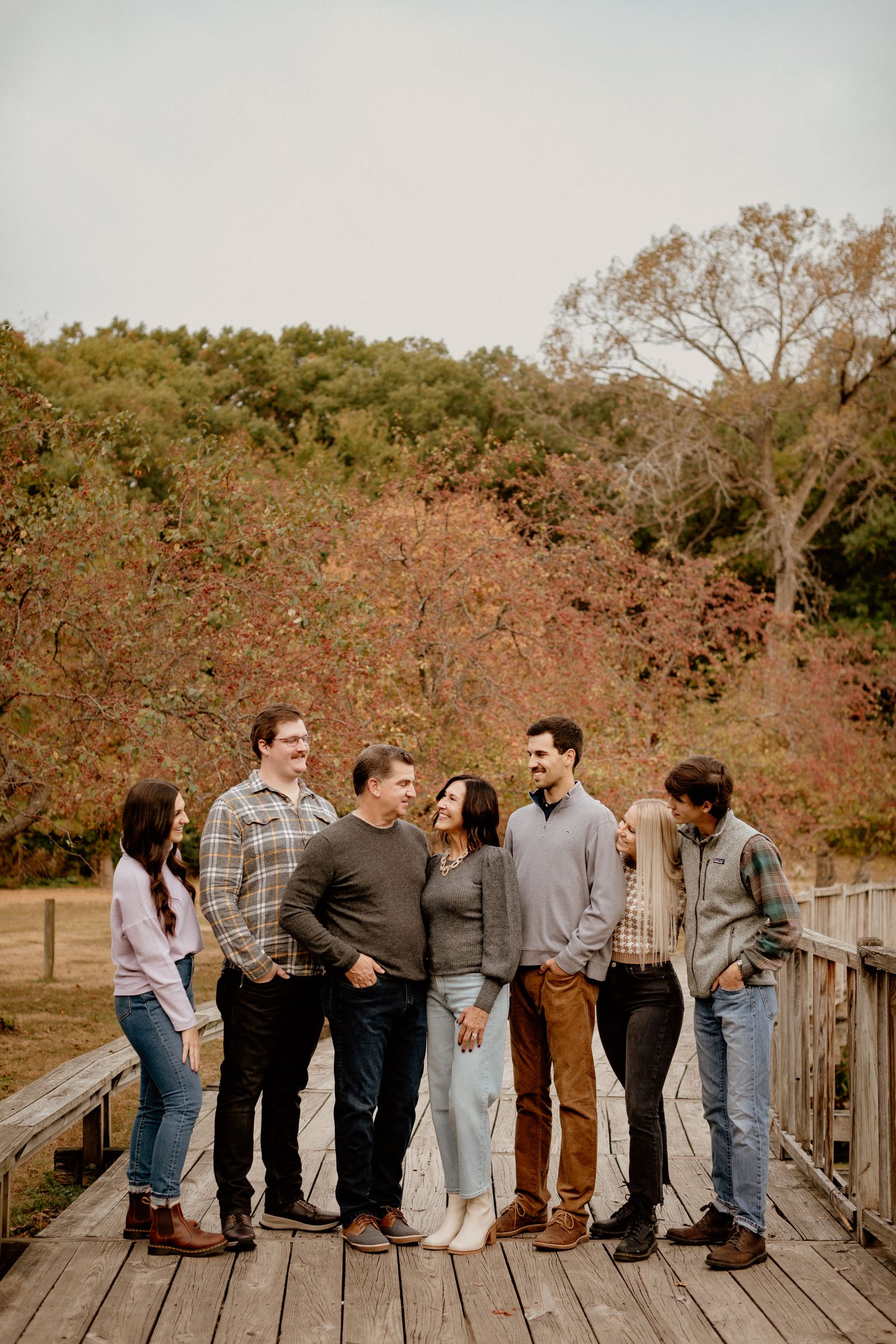 A group of seven young adults laughing together on a wooden bridge surrounded by autumn foliage.