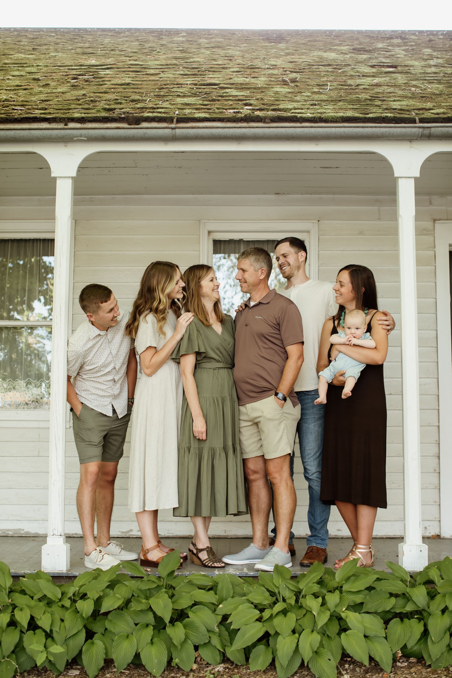 A joyful family of seven poses on a white farmhouse porch, laughing together, one woman holding a baby.