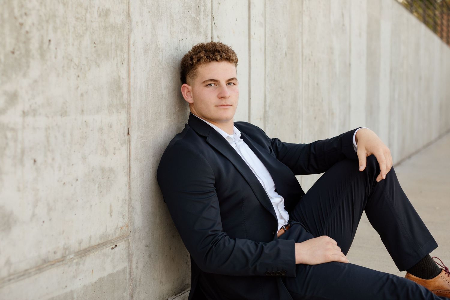 Young man in a navy suit sitting relaxed against a concrete wall outdoors.