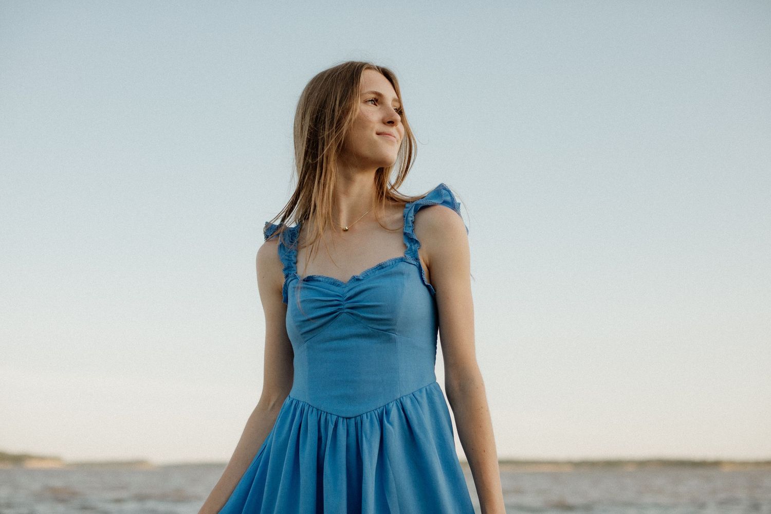 Young woman in a blue sundress standing outdoors near water, looking away with a serene expression.