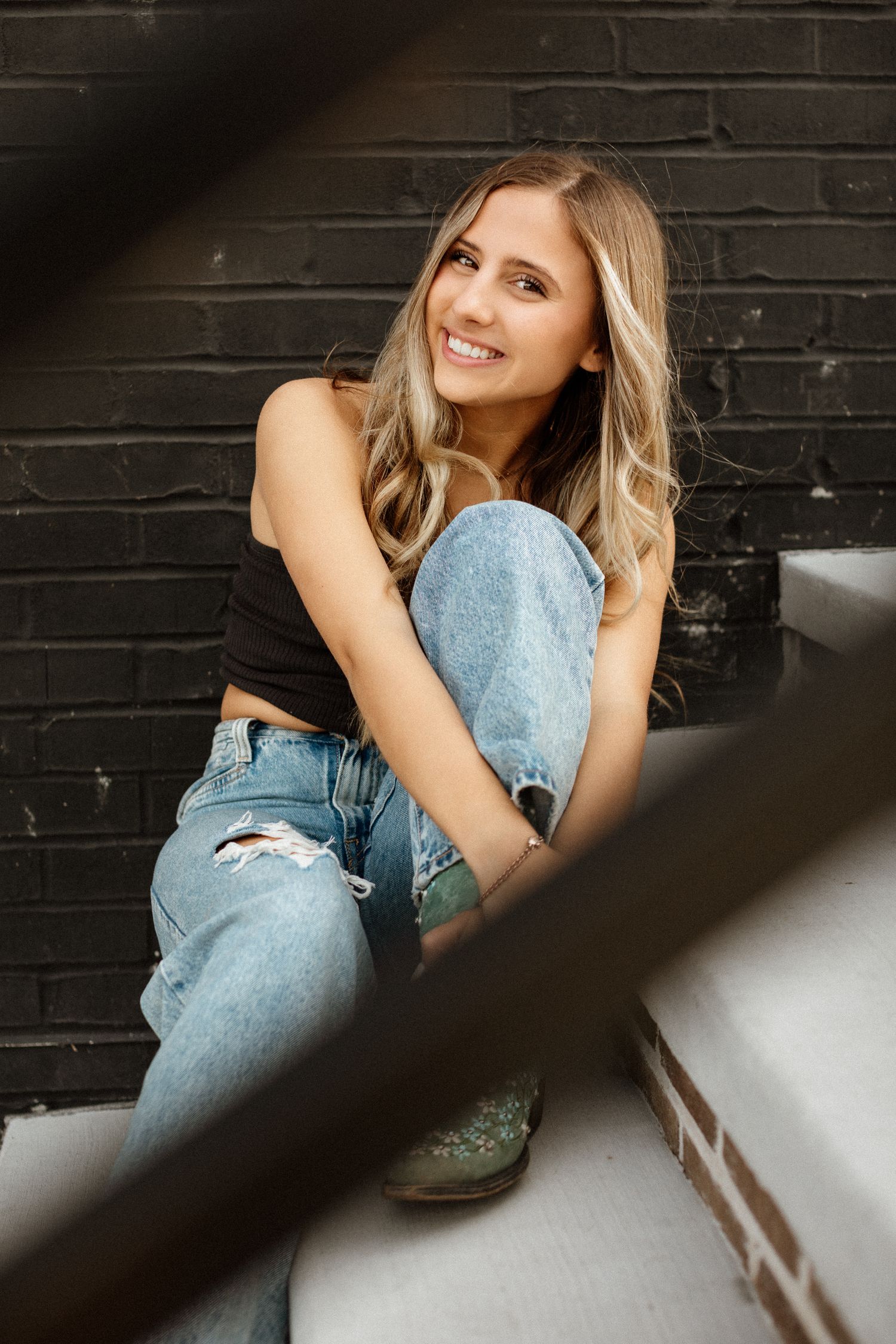 Smiling blonde woman in black crop top and ripped jeans sitting on stairs against a brick wall.