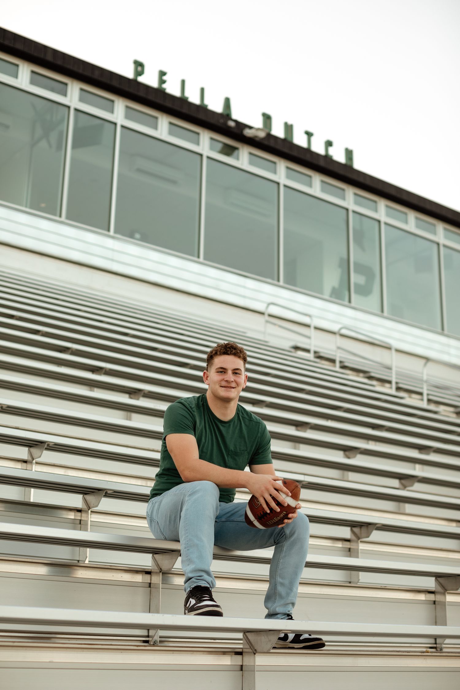 Smiling teen in green shirt holding a football while sitting on stadium bleachers at Pella Dutch.