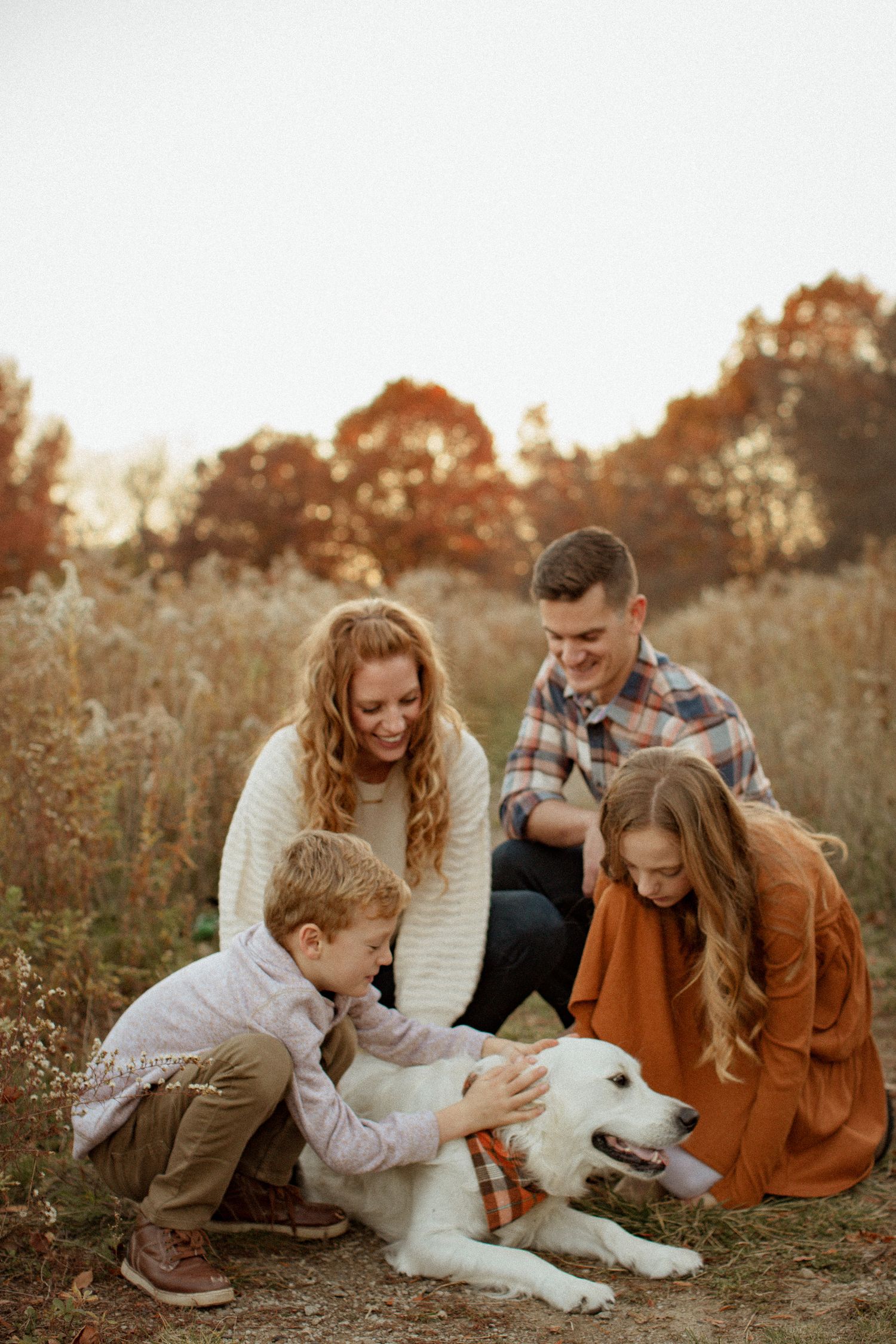 A happy family with two children and a white dog pose together in an autumn field at golden hour.