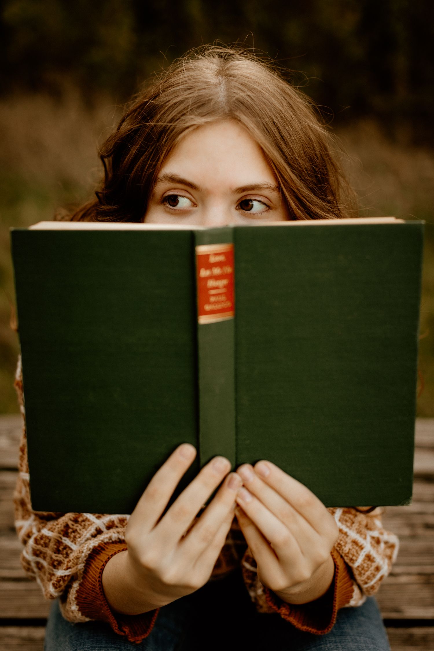 Young woman peeking over a green hardcover book with curious eyes outdoors.