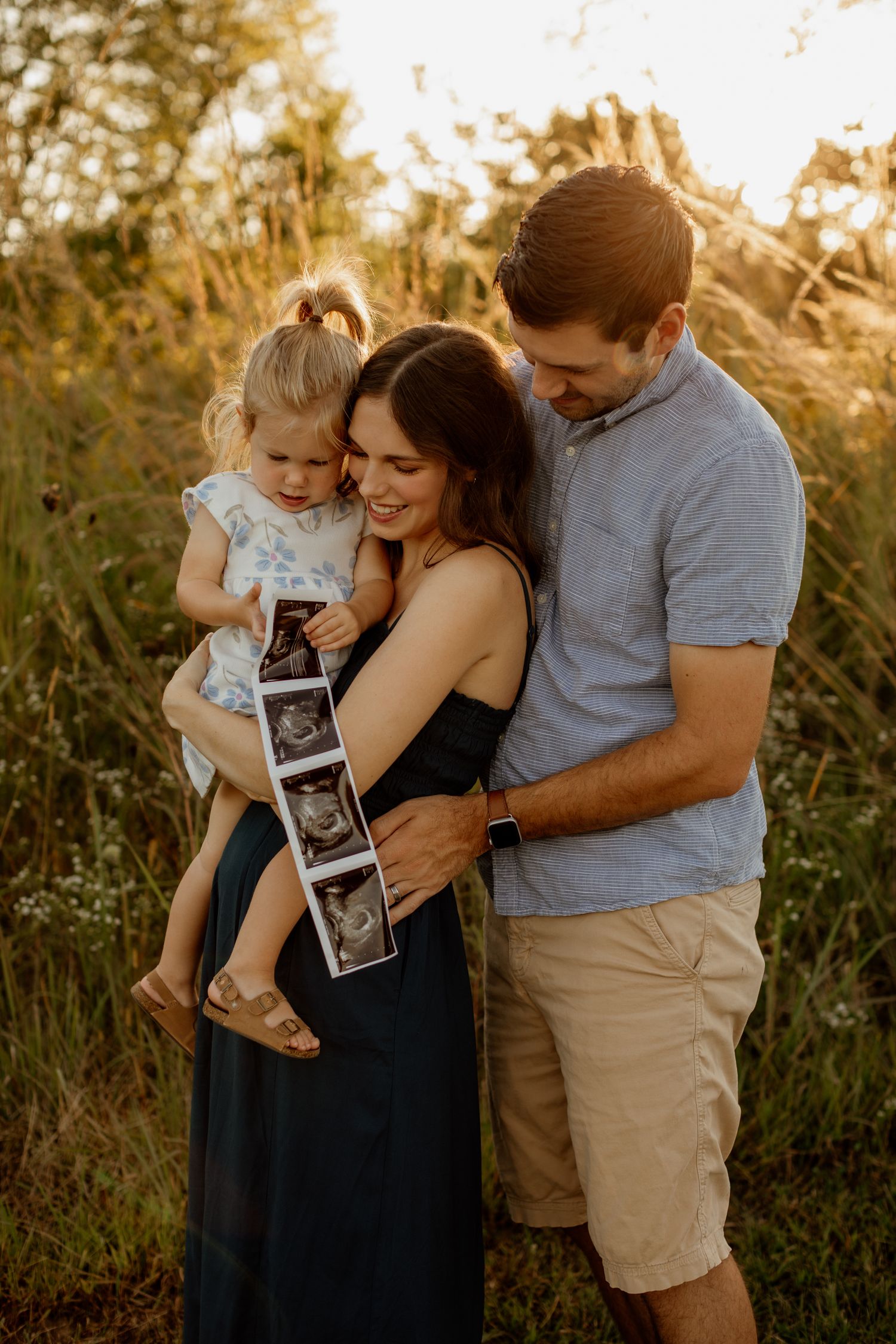Family of three holding ultrasound photos together in a warm sunset field setting.