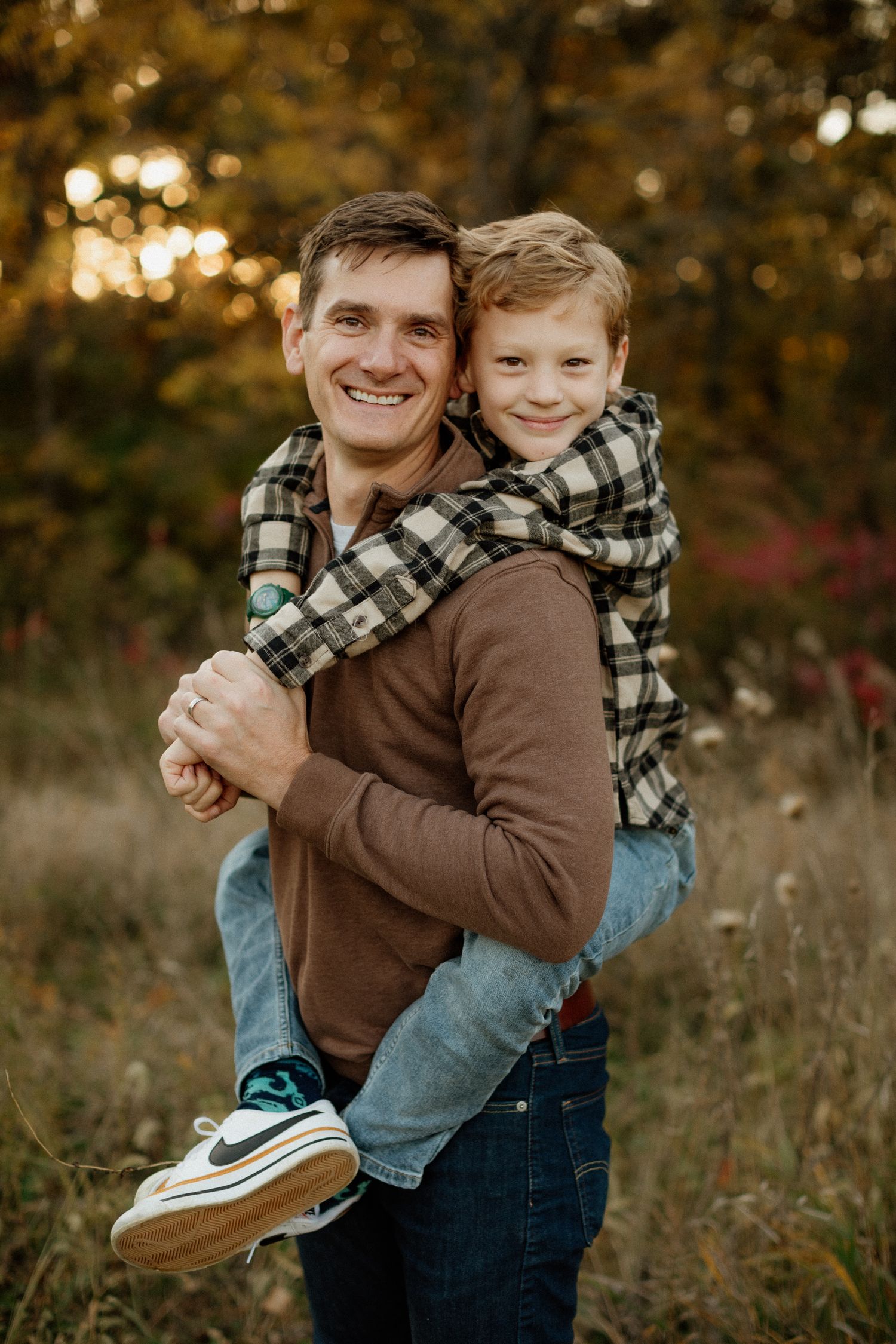 A smiling man gives a young red-haired boy a piggyback ride in an autumn field.