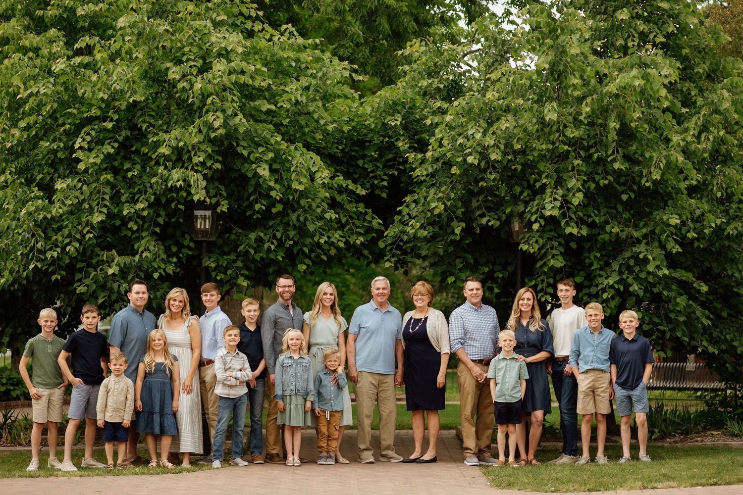 Large multigenerational family posing together outdoors in front of lush green trees on a sunny day.