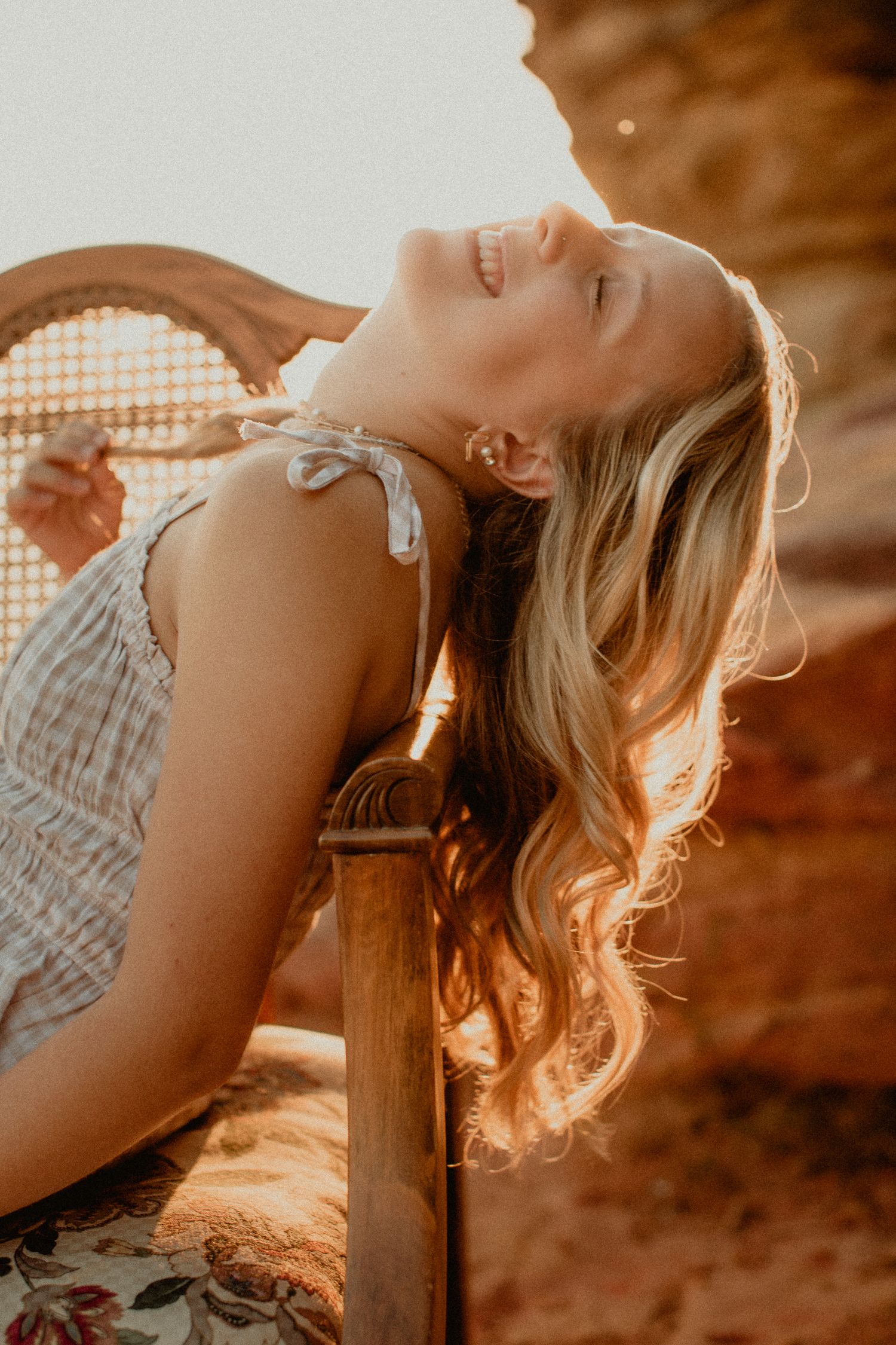Blonde woman laughing with head tilted back in golden sunset light beside wooden chair.