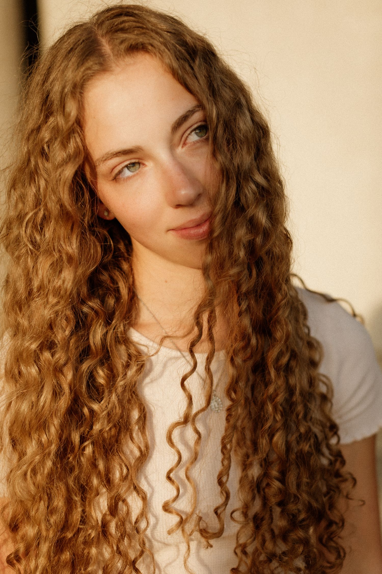 Young woman with long curly auburn hair gazes softly, wearing a white t-shirt in golden hour light.