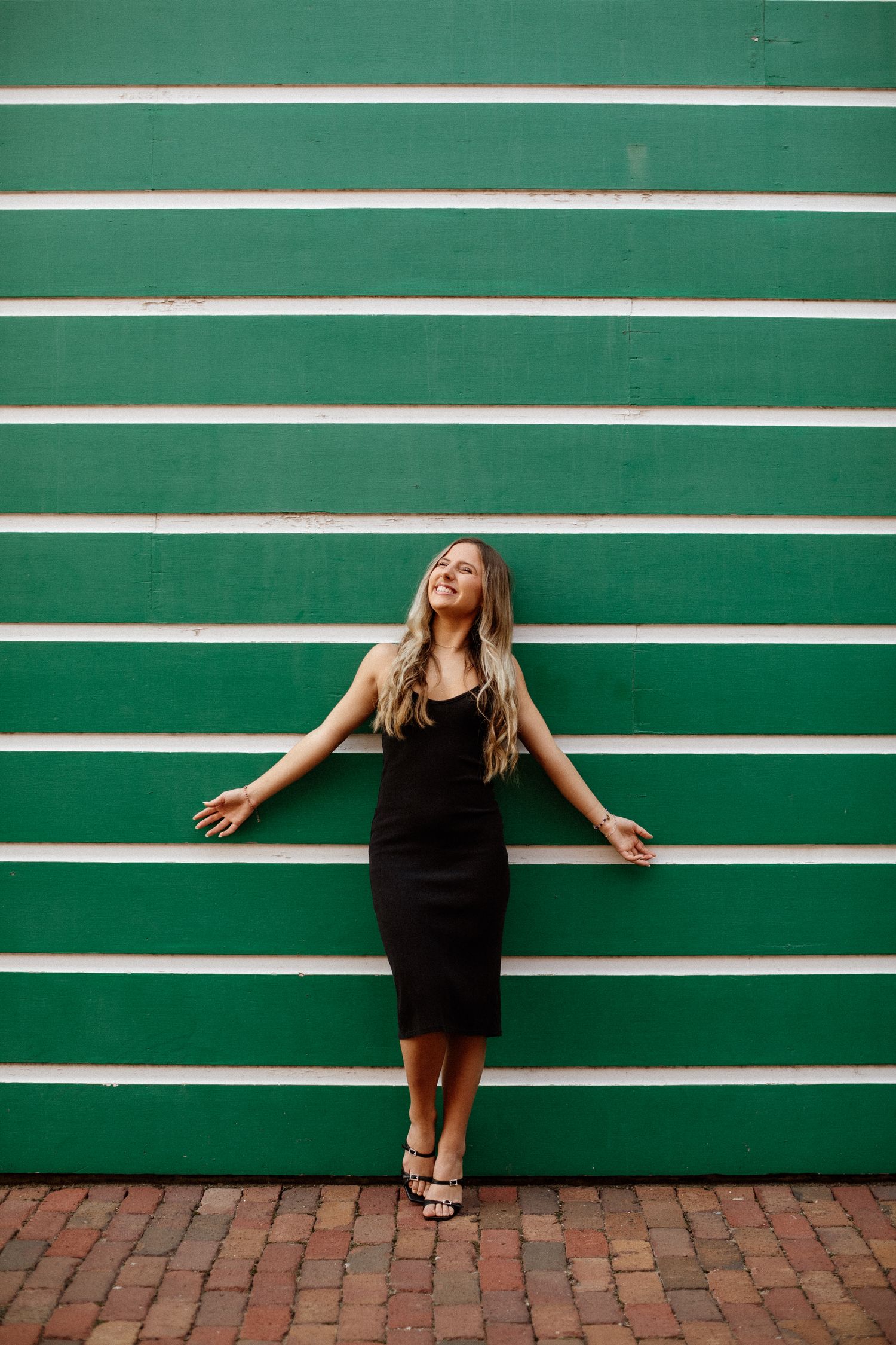 Blonde woman in black sleeveless dress posing with arms outstretched against a green and white striped wall.