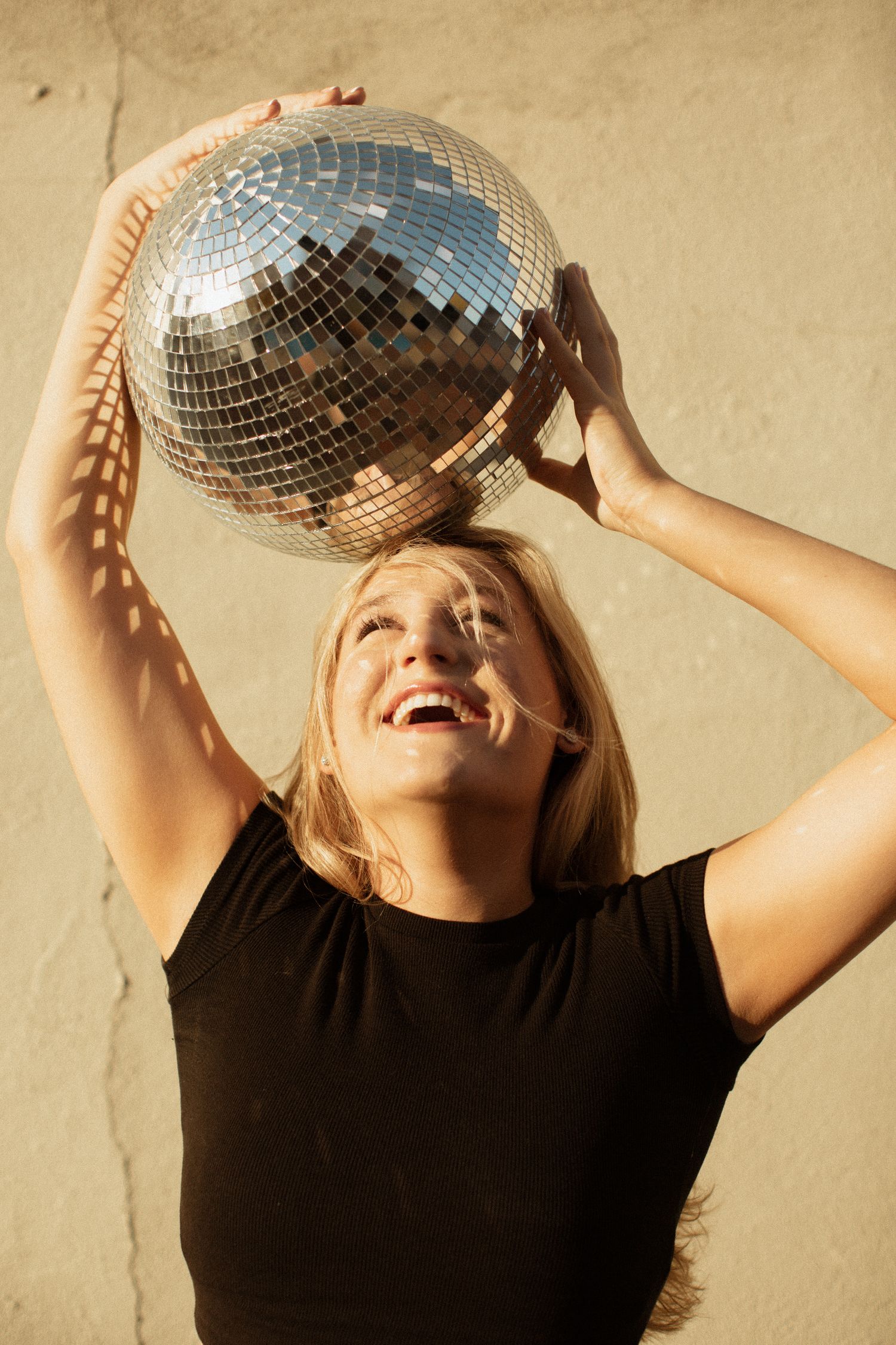Laughing blonde woman in black tee holding a shiny disco ball above her head against a beige wall.
