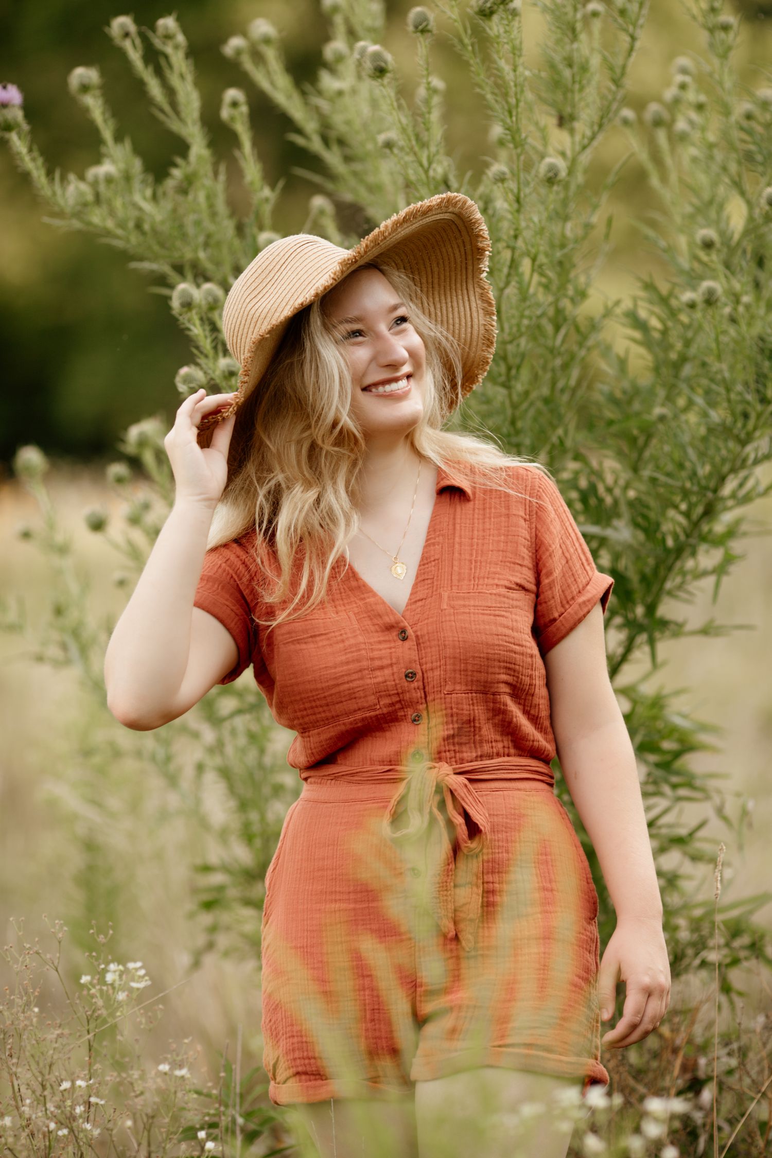 Smiling blonde woman in orange romper holds a straw hat while standing among wildflowers in a field.