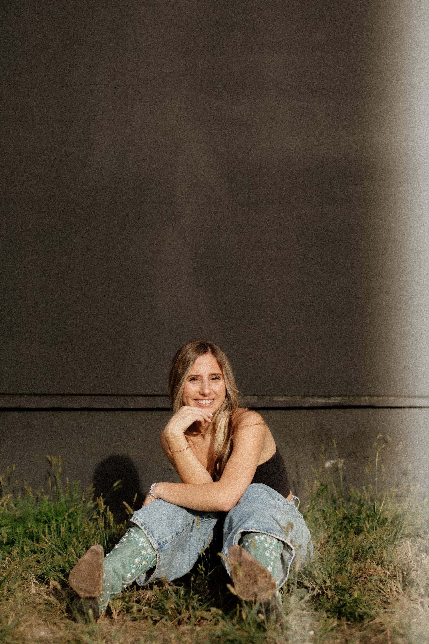 Smiling young woman in black top and ripped jeans sitting cross-legged on grass against a dark wall.