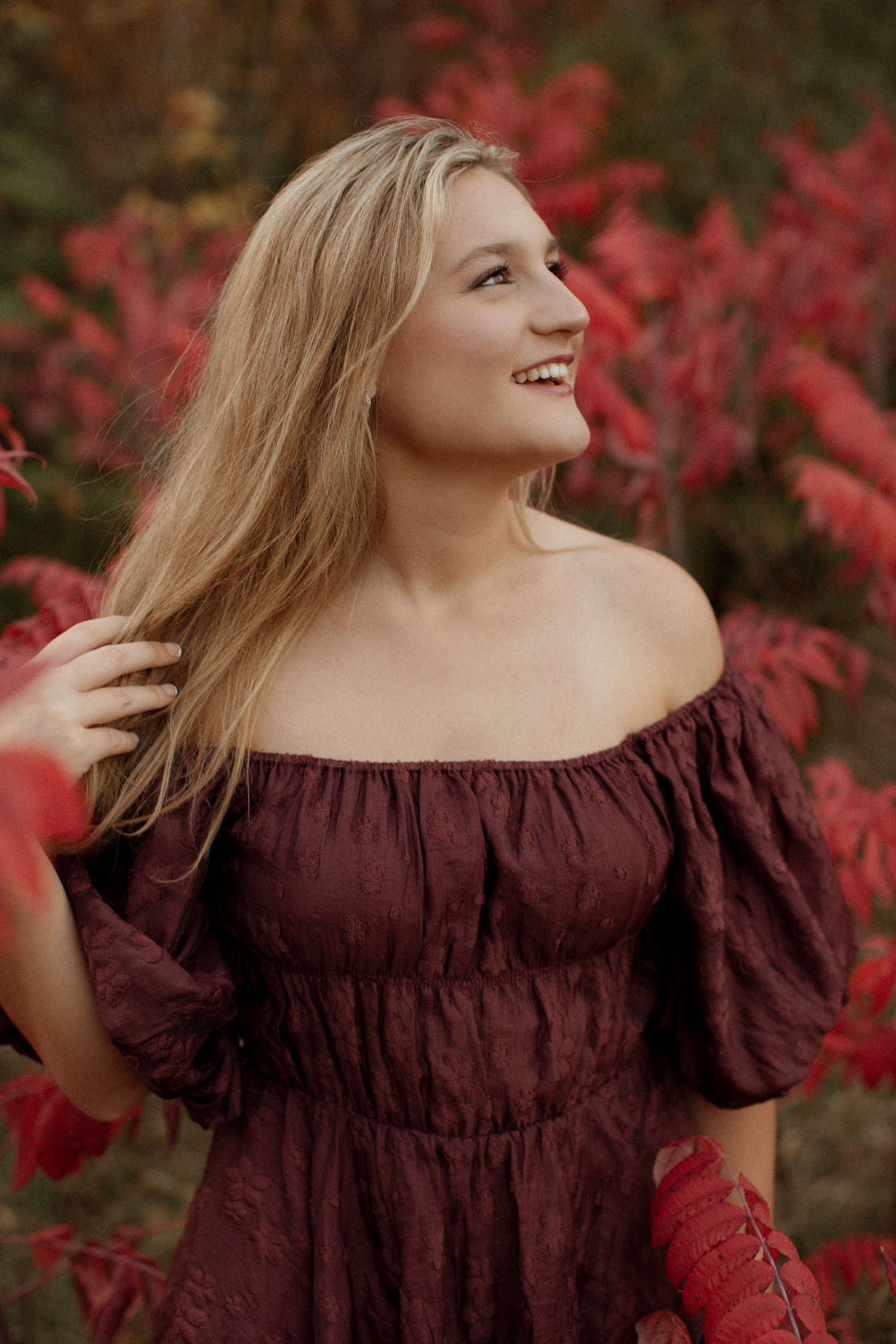 Smiling blonde woman in a burgundy off-shoulder dress posing near vibrant red autumn foliage.