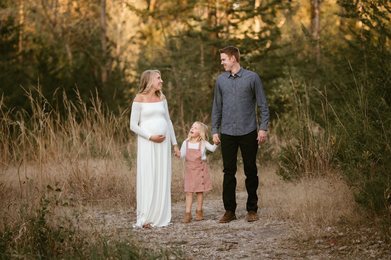 Pregnant woman in white gown walks with toddler and man on forest path during golden hour maternity photo session.