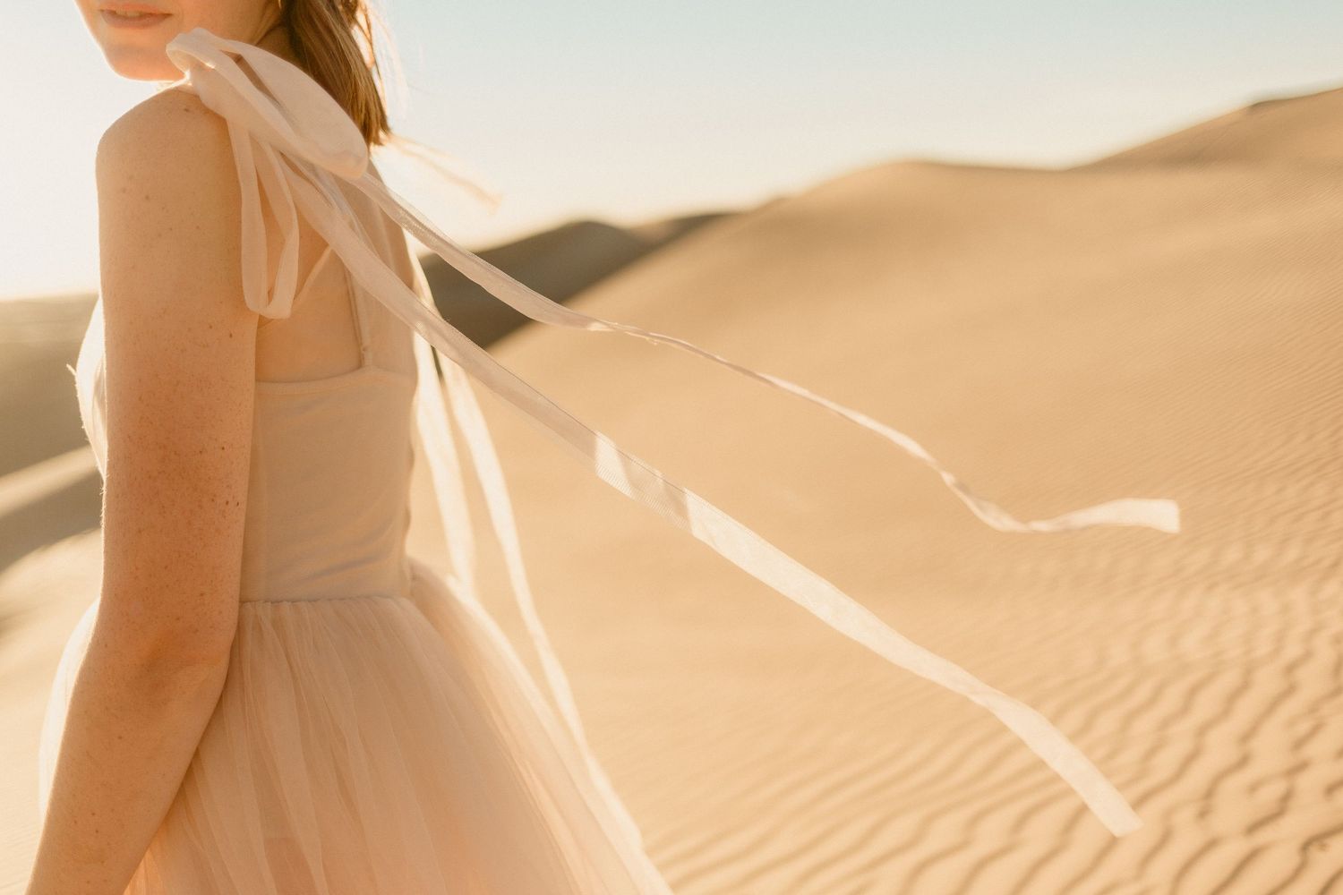 Woman in a blush tulle dress holding flowing ribbons in a sunlit sand dune.