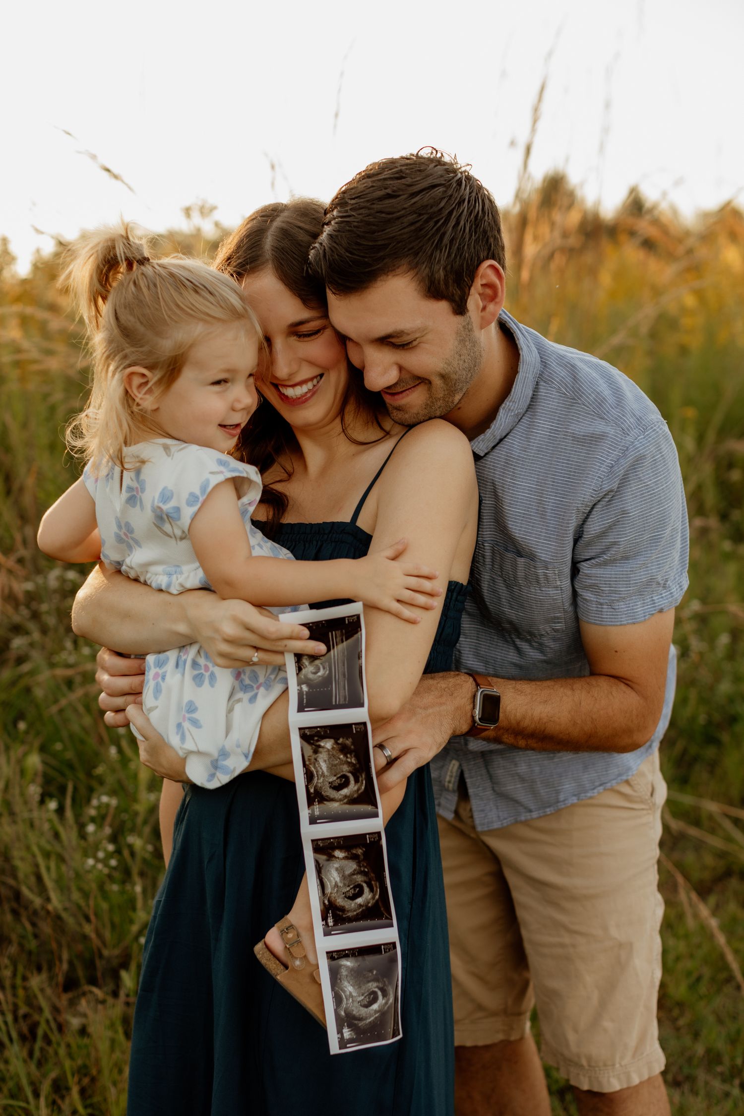 Smiling couple with young daughter holding ultrasound images in a sunny meadow.