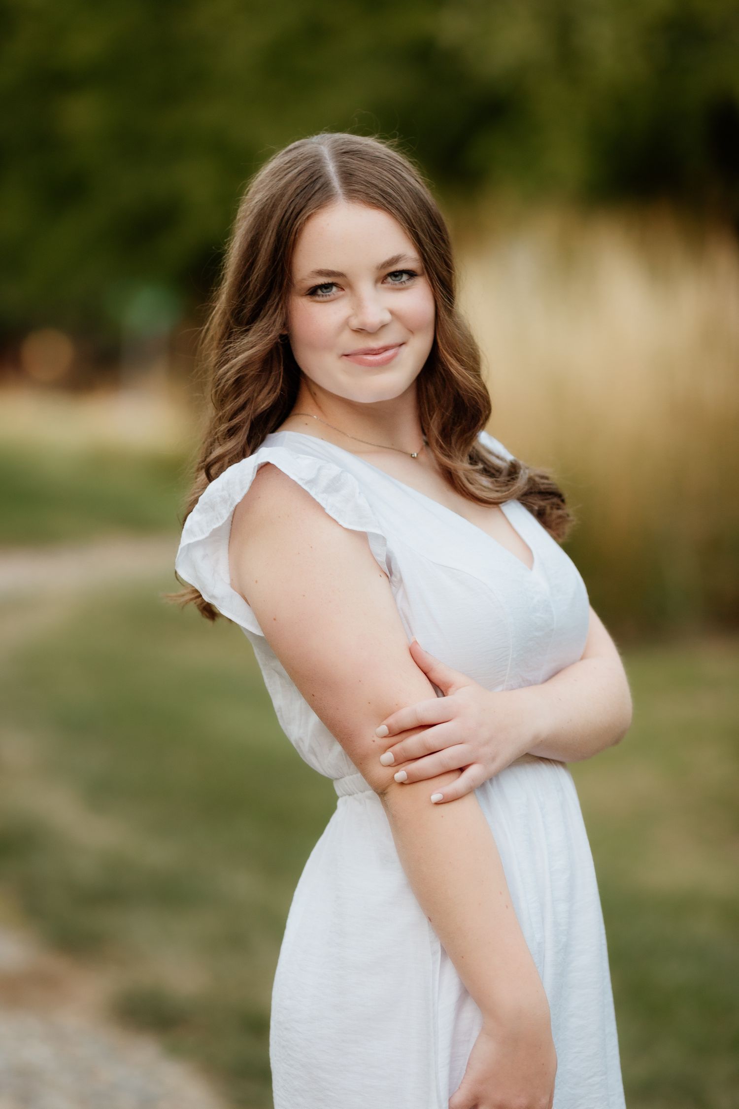 Smiling young woman in a white dress with wavy brown hair posing outdoors in a green field.