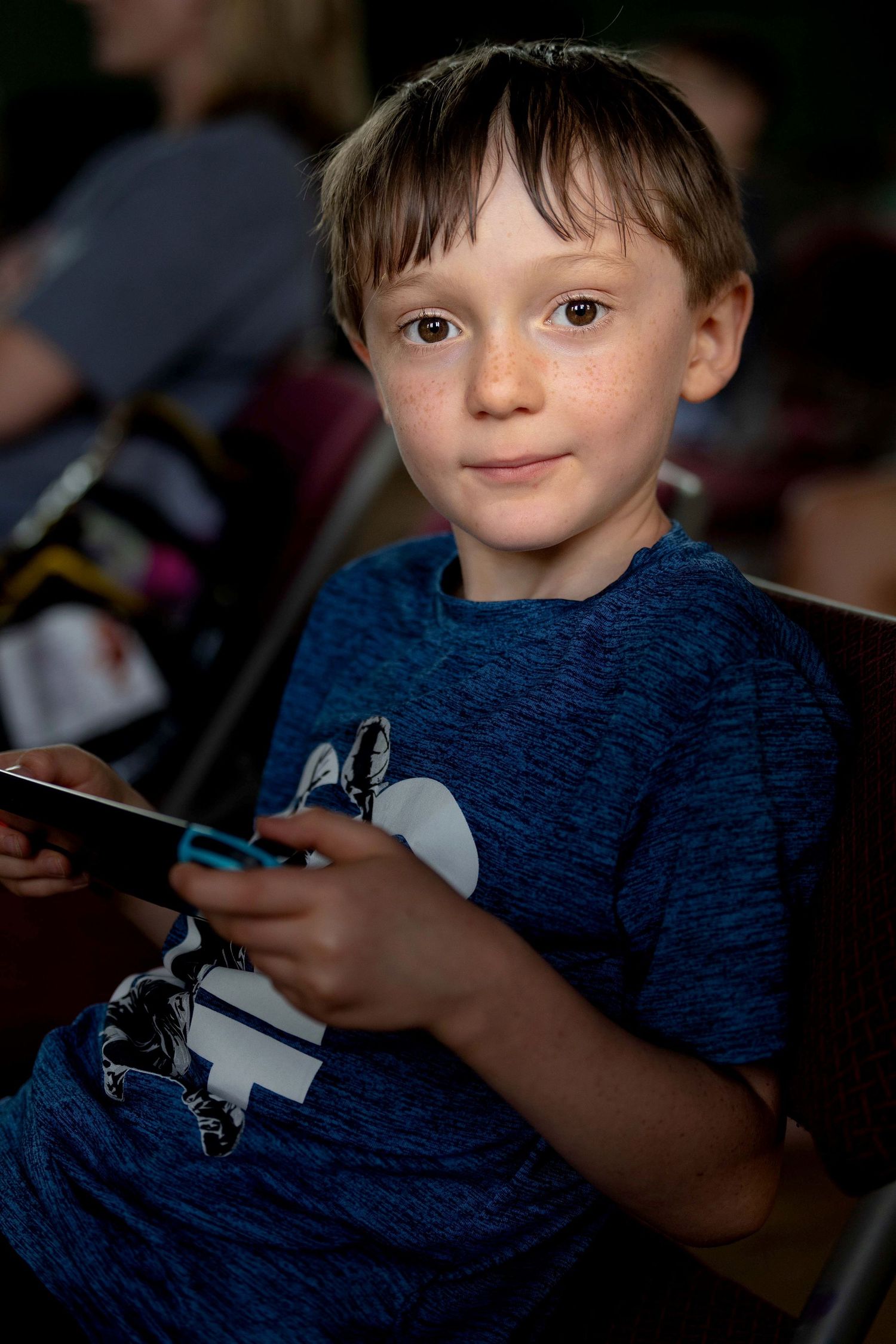 A young freckled boy in a blue t-shirt holds a Nintendo Switch controller while seated in an audience.