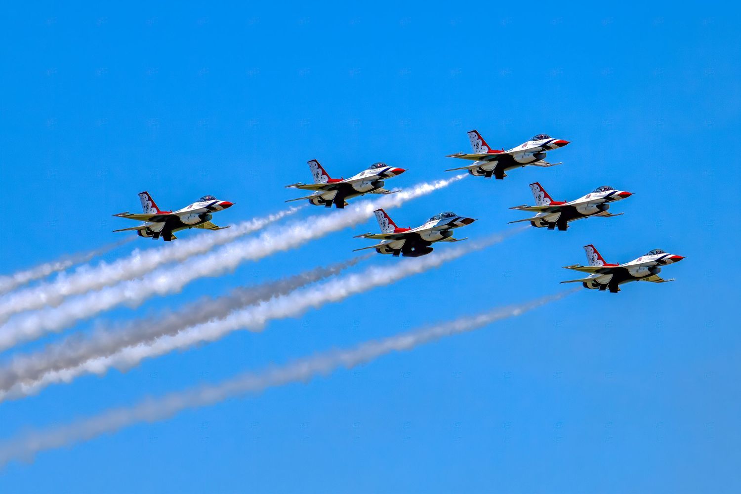 Six US Air Force Thunderbirds jets fly in tight diamond formation leaving white smoke trails against a clear blue sky.