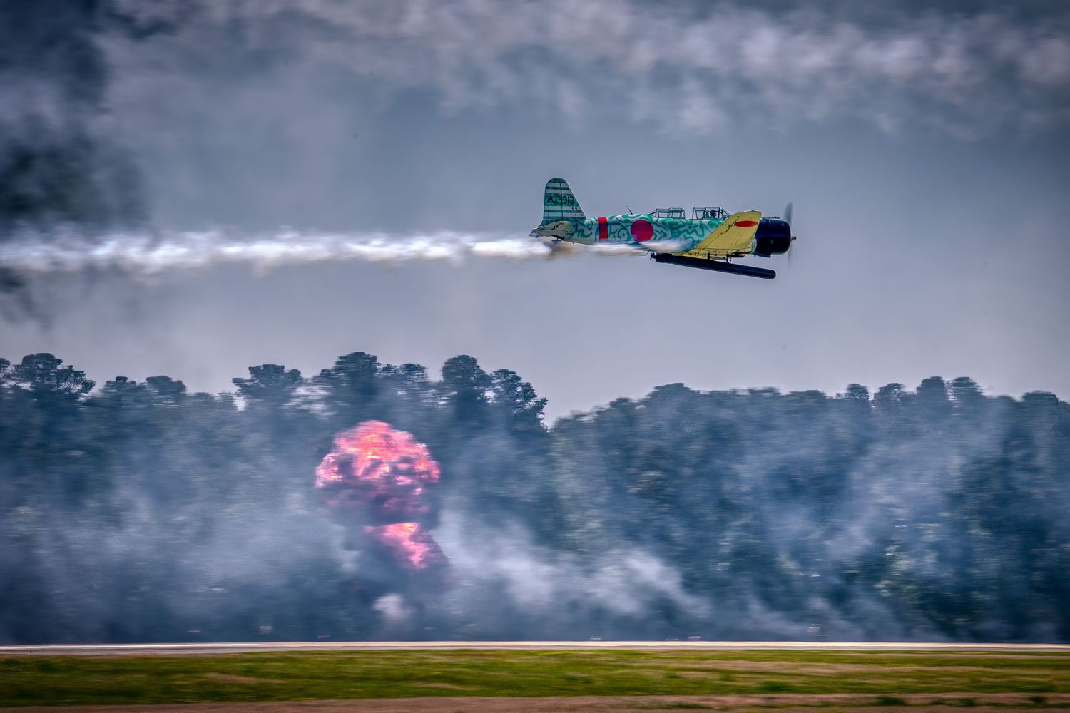 Colorful stunt plane flying low over airfield with dramatic pink smoke explosion on ground.