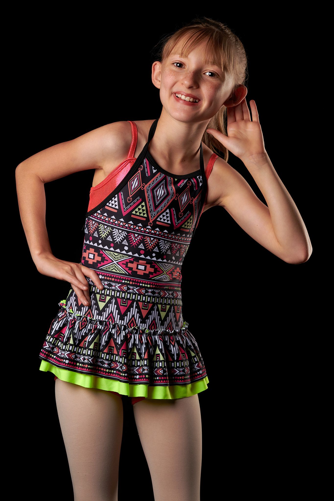 Smiling preteen girl in colorful geometric dance costume posing against black background.