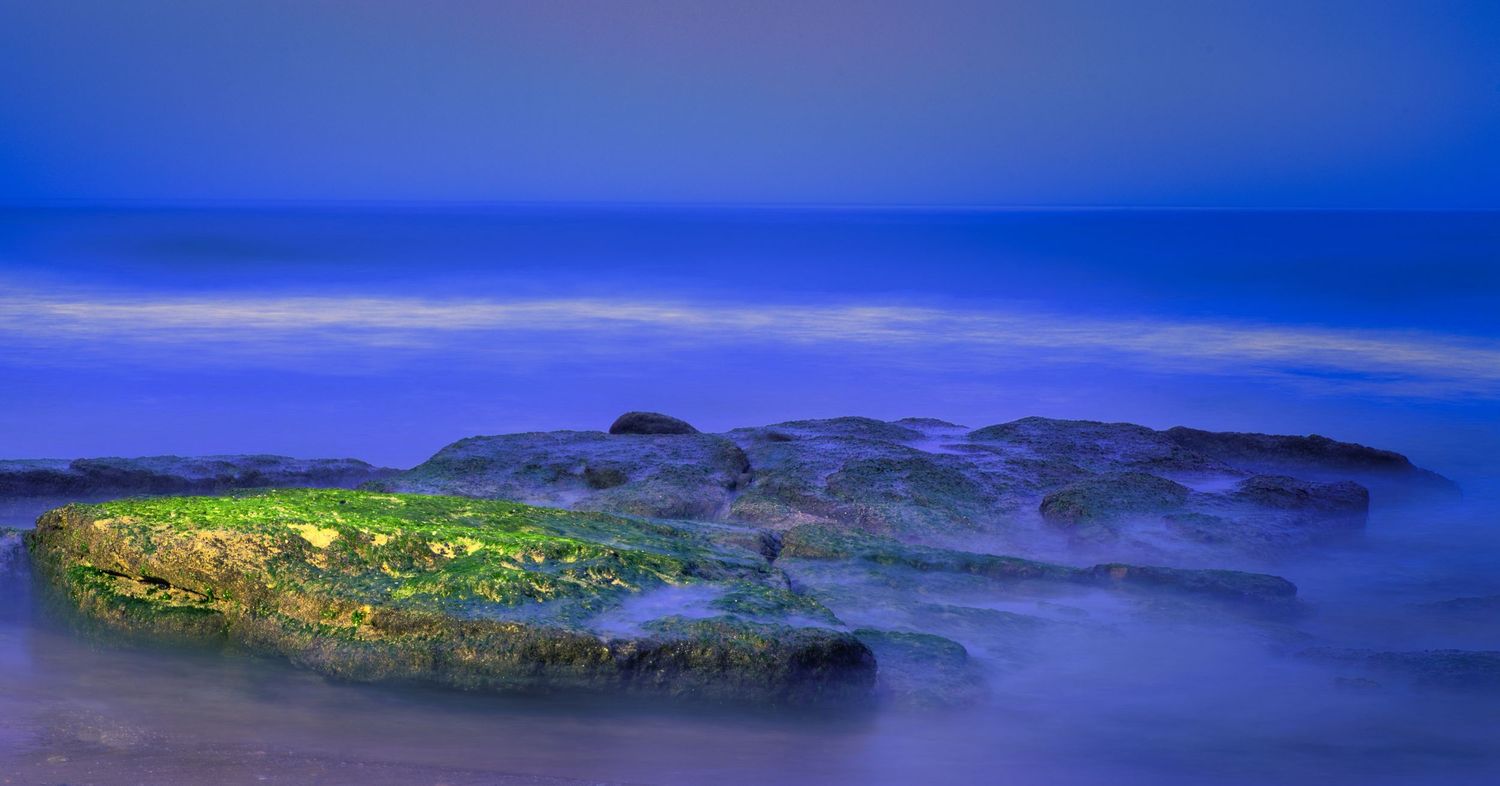 Mossy rocks surrounded by misty blue ocean waves at dusk.