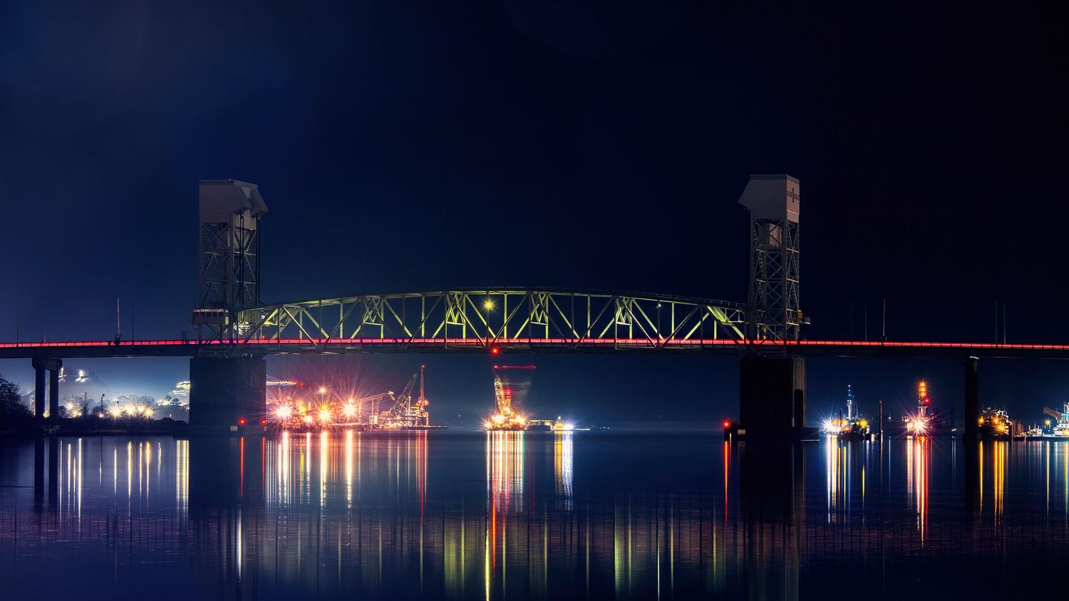 Illuminated bridge reflecting colorful lights on calm water at night.