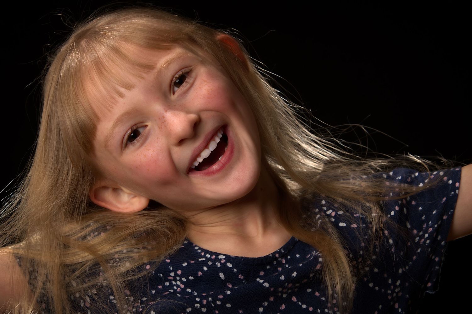 Joyful young blonde girl laughing with hair flowing against a dark background.