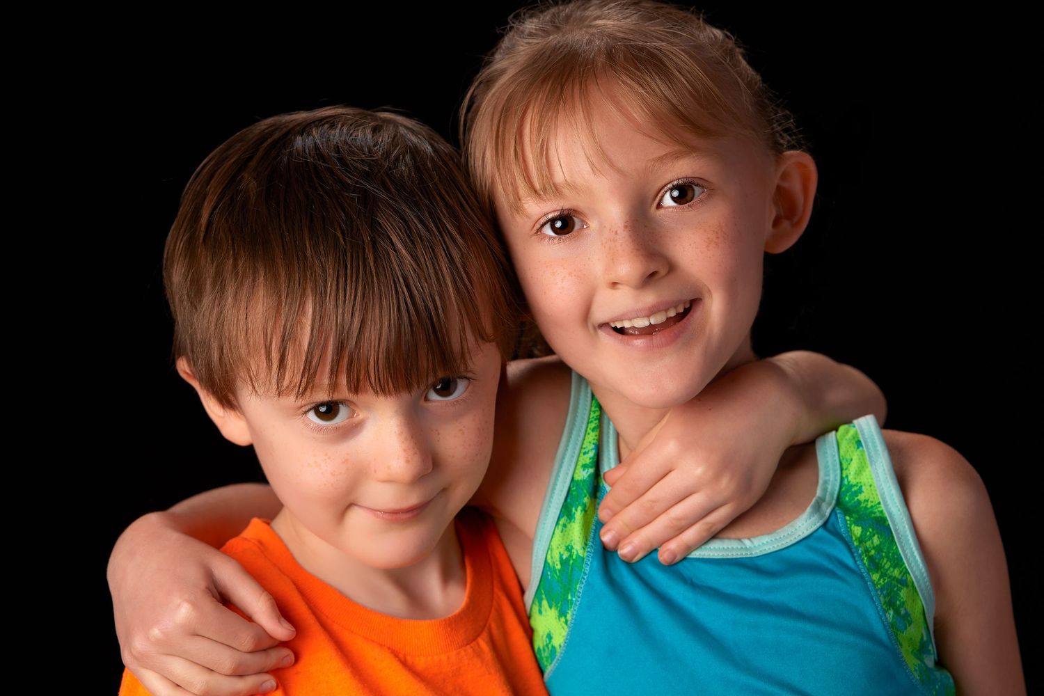 Two young children, a boy in orange and a girl in blue, smile and hug against a black background.