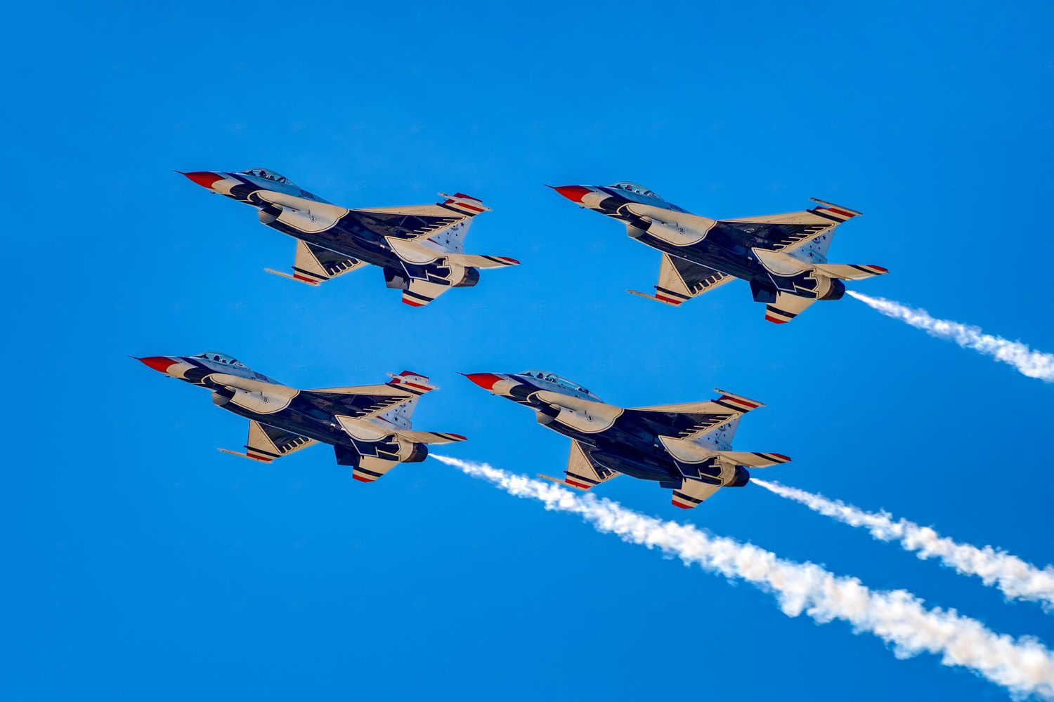 Four US Air Force Thunderbirds F-16 jets fly in precise diamond formation with white contrails on a clear blue sky.
