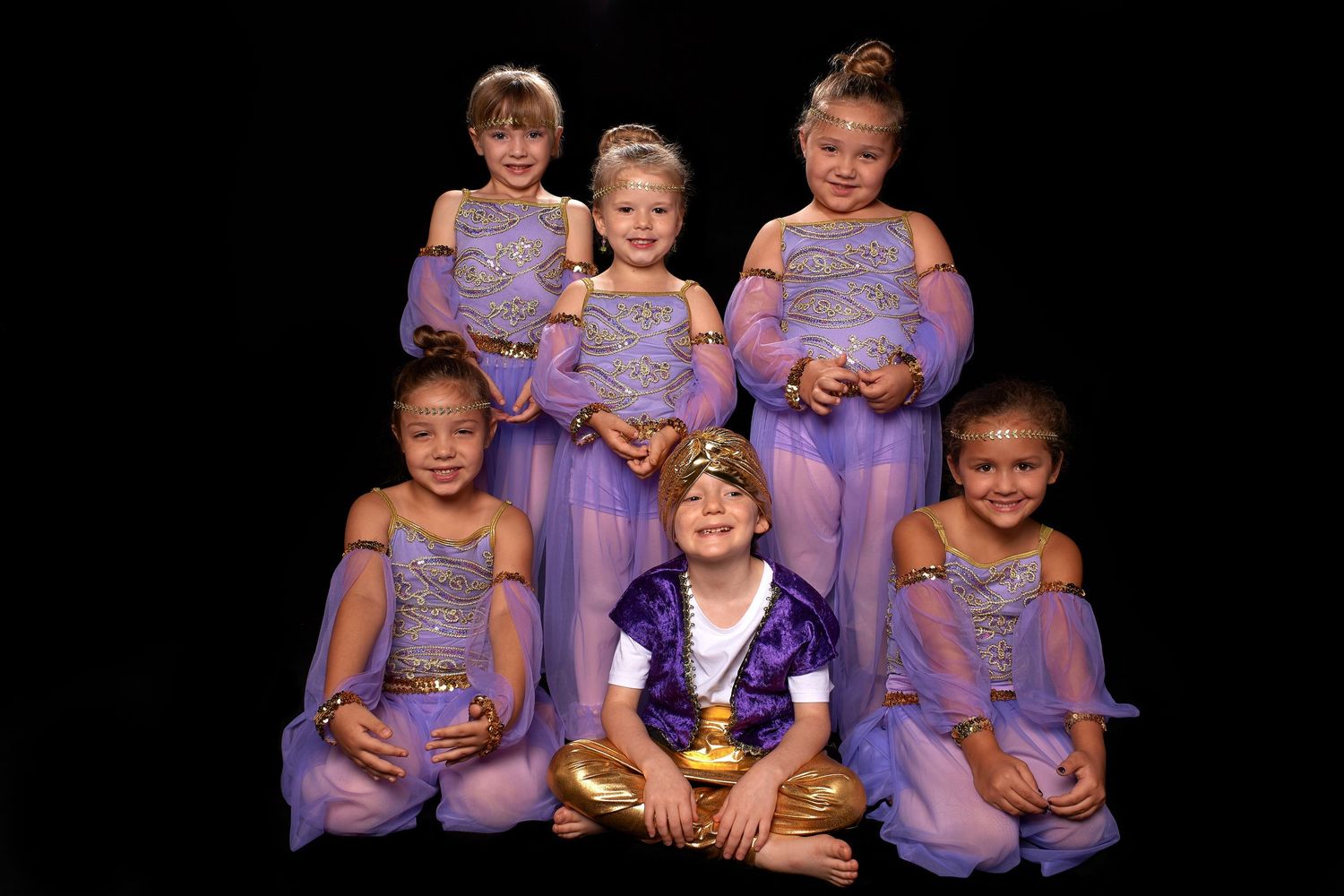 Group of six young girls in purple dance costumes posing together against black background.