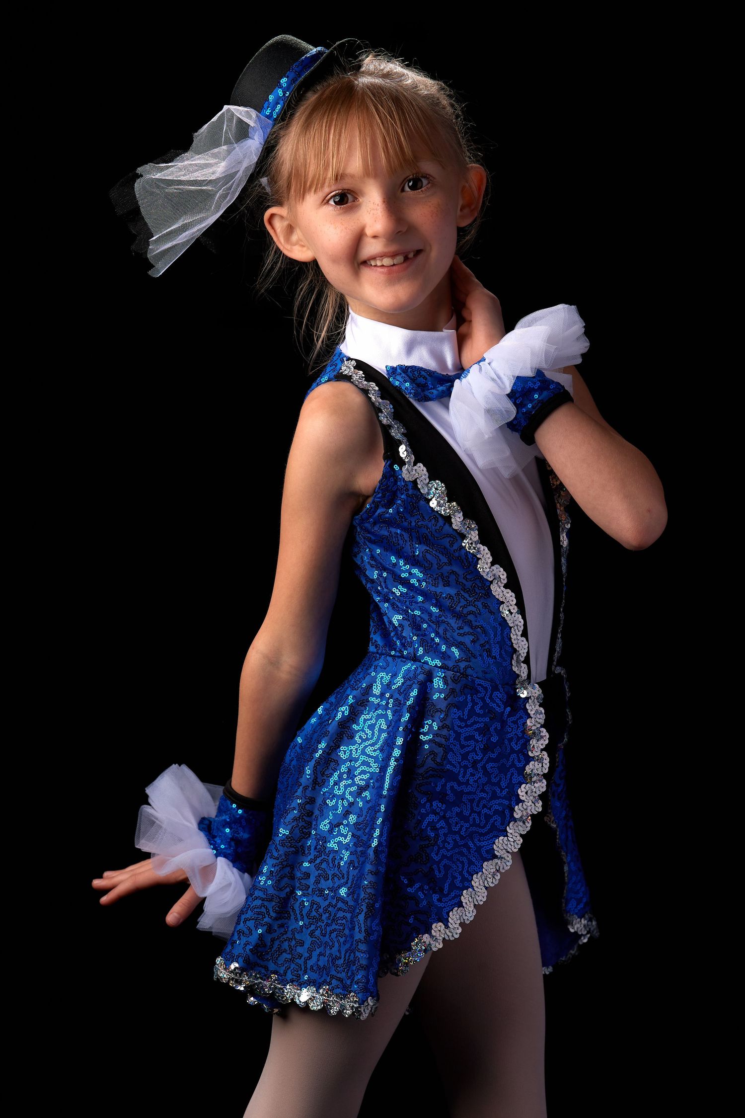 Young girl in blue sequin dance costume with hat posing against black background.