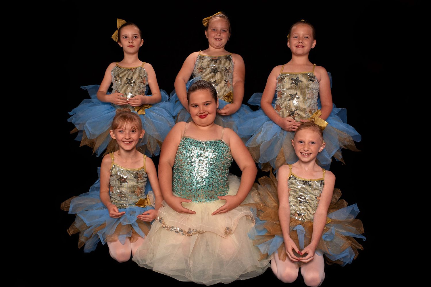 Six young girls in sparkly blue and silver ballet tutus pose together for a dance recital group photo.