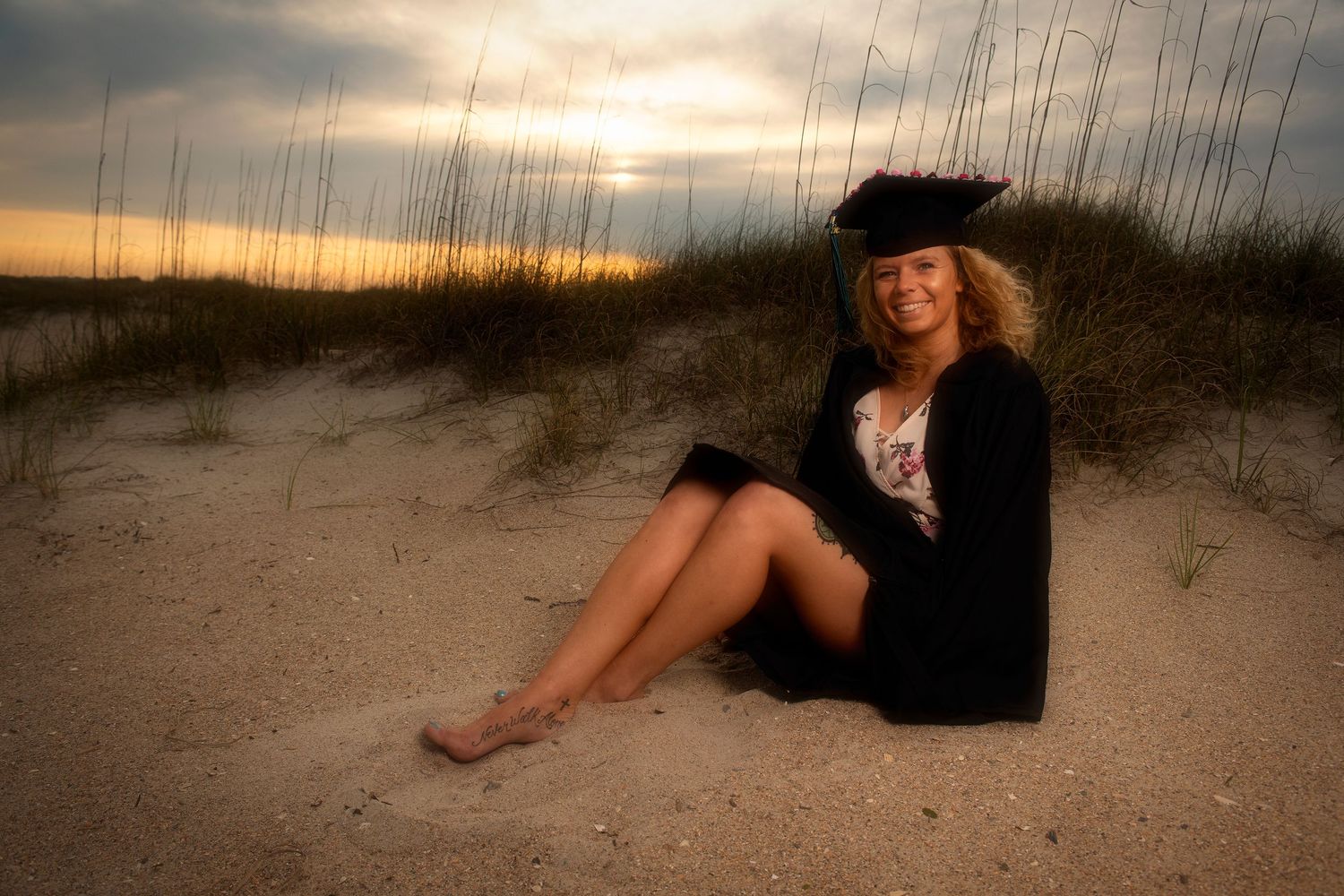 Female graduate in cap and gown sitting barefoot on sandy beach dunes at sunset.
