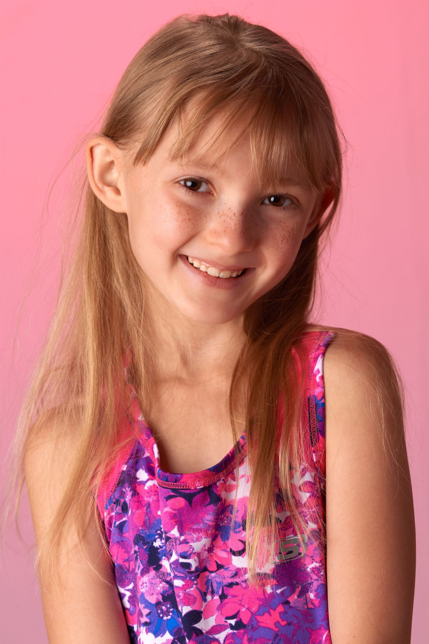 Smiling young girl with long blonde hair and freckles wearing a pink floral dress against a pink background.