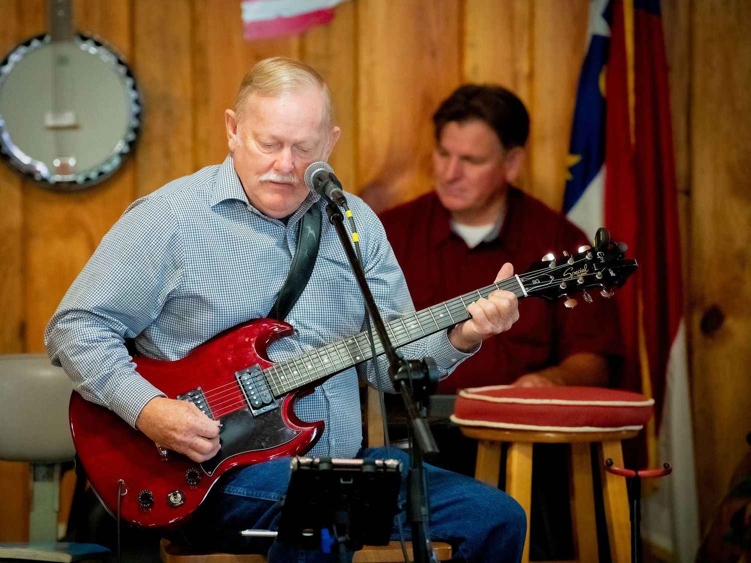 Older man playing red electric guitar at microphone with another musician in background.