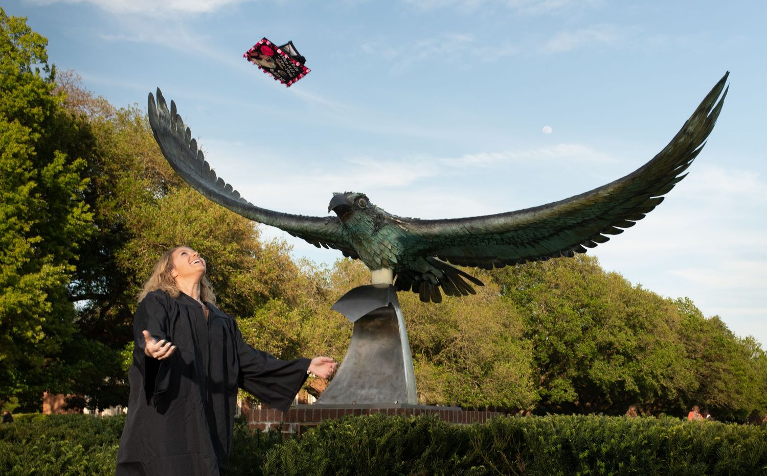 Female graduate in black gown tossing decorated cap near large bird statue on campus.