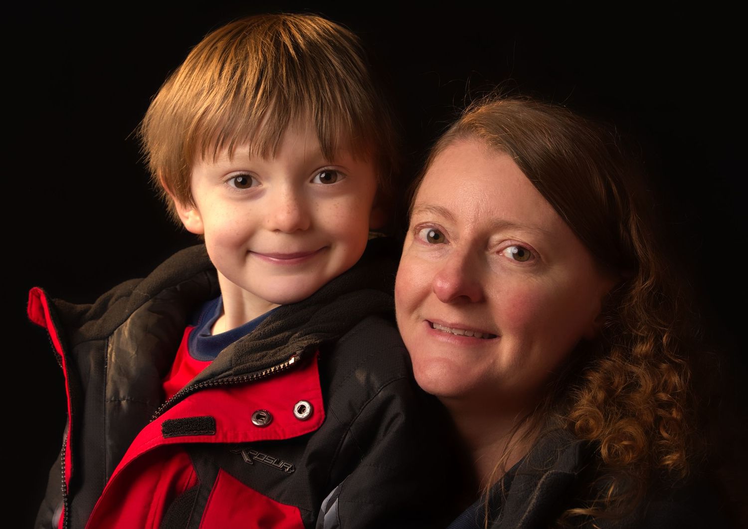 Portrait of a young red-haired boy and his smiling mother posed together against a dark background.