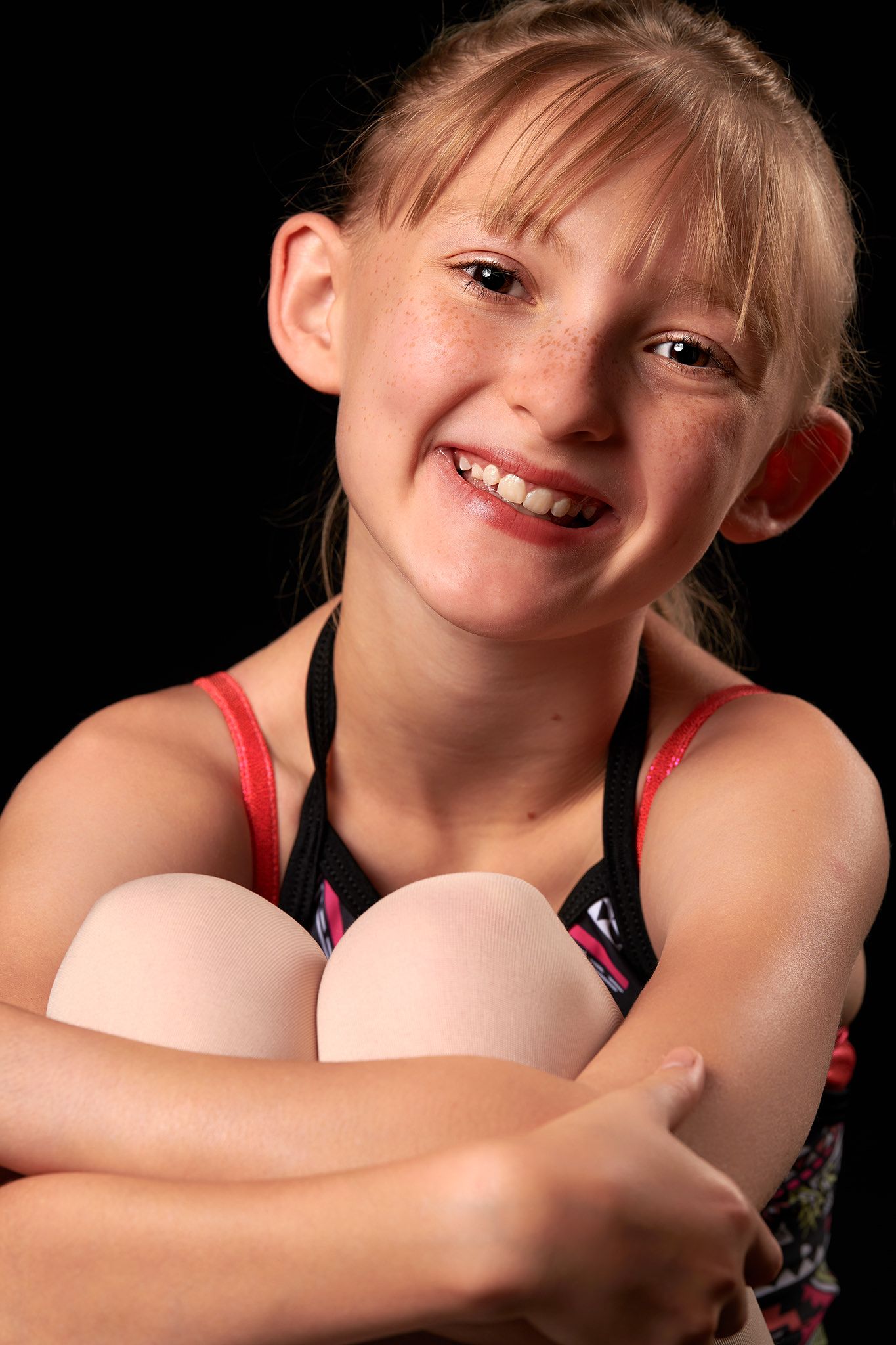 Happy freckled blonde girl hugging her knees and smiling in a studio portrait.
