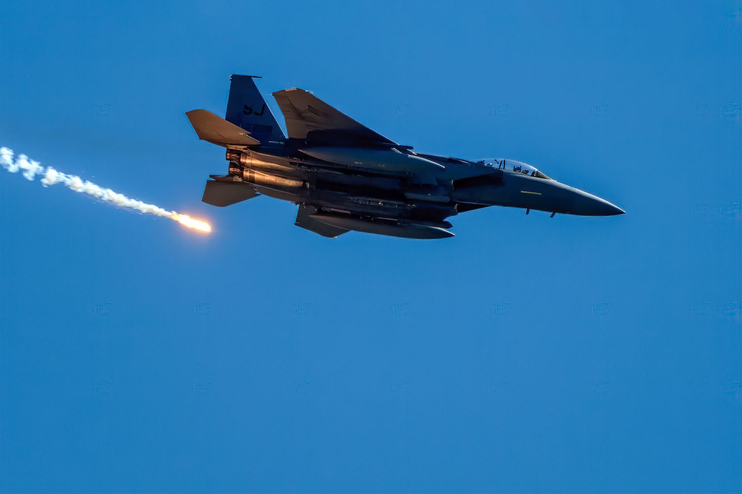 An F-15 fighter jet releases bright flares while banking sharply against a deep blue twilight sky.
