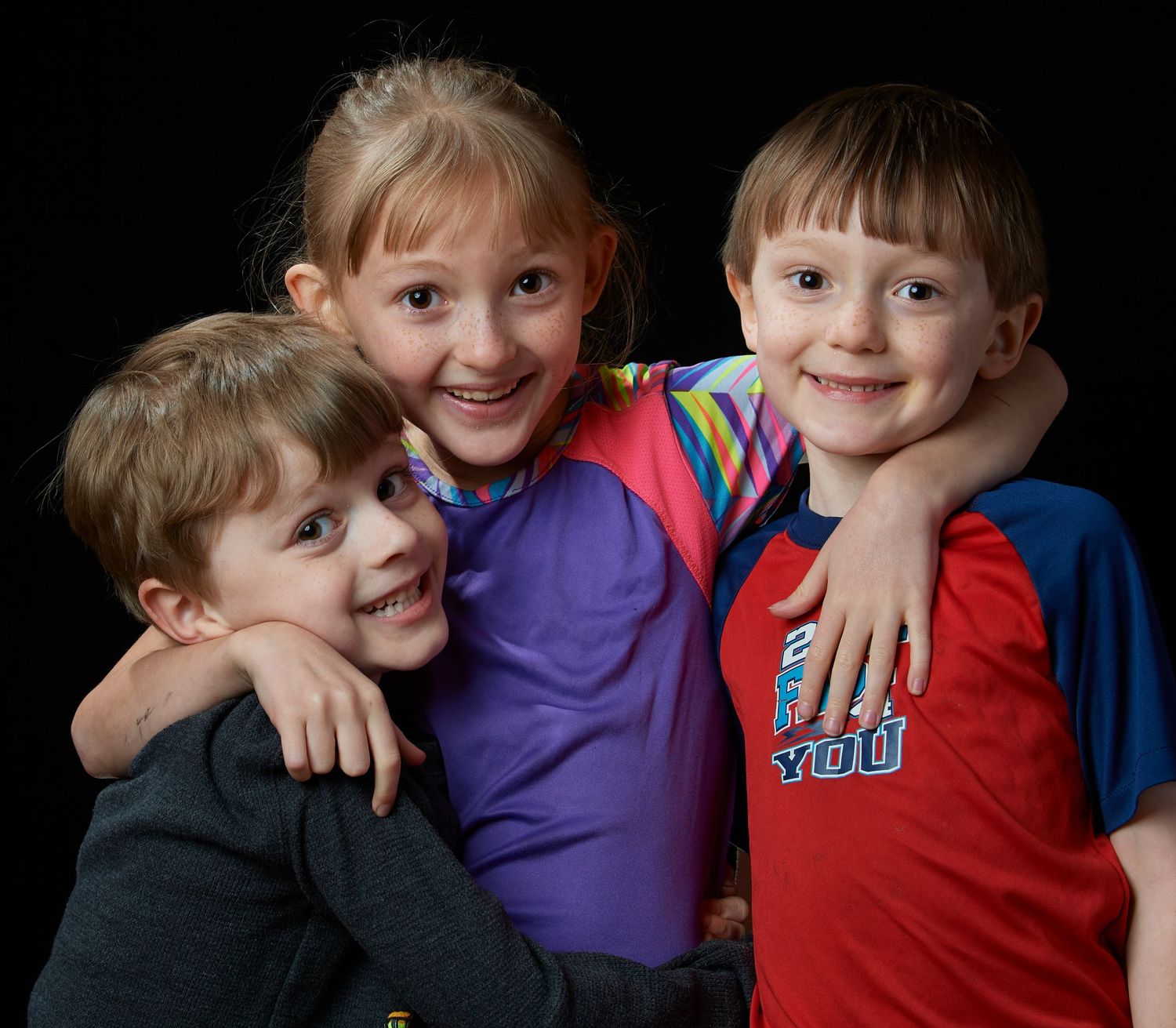 Three happy children hugging each other and smiling in a studio portrait with black background.