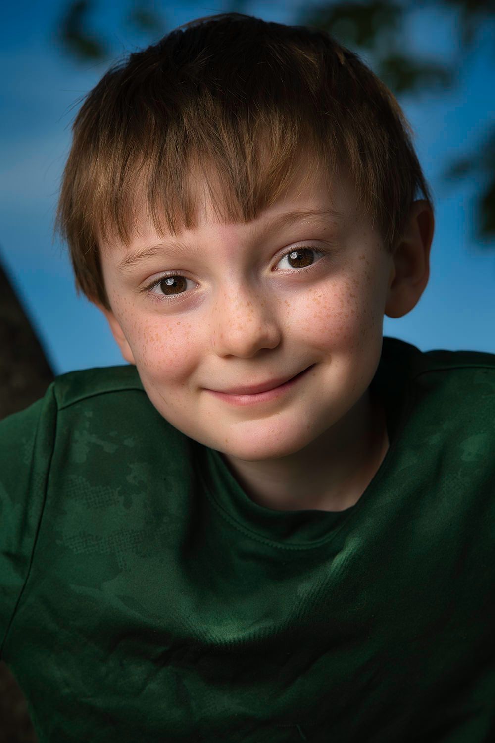 A smiling red-haired freckled boy in a green t-shirt poses outdoors against a blue sky background.
