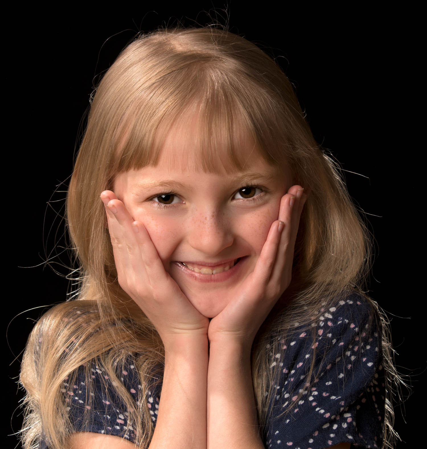 Smiling young blonde girl posing with hands on cheeks against a black background.