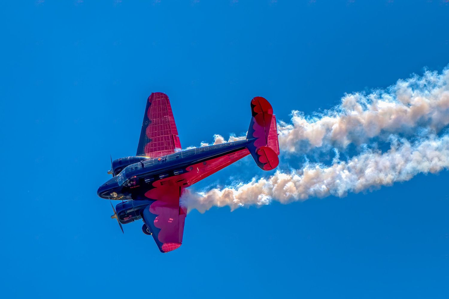 Red and blue vintage aircraft performing aerobatics against clear blue sky with white smoke trail.