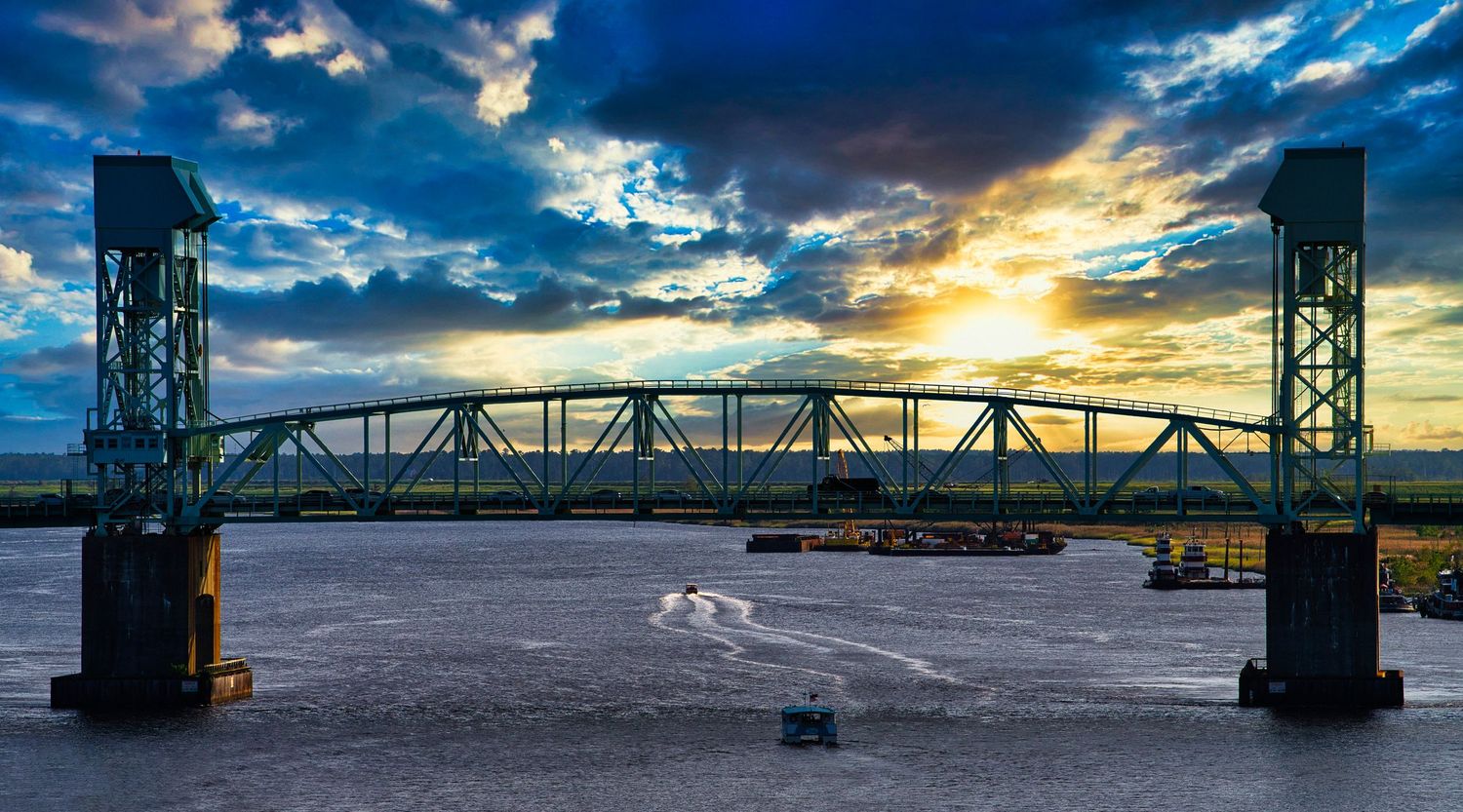 A steel lift bridge spans a wide river at sunset with dramatic clouds and a small boat passing beneath.