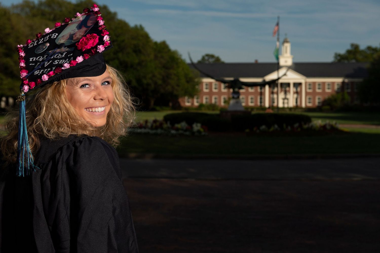 Smiling female graduate in cap and gown standing on a college campus lawn at golden hour.