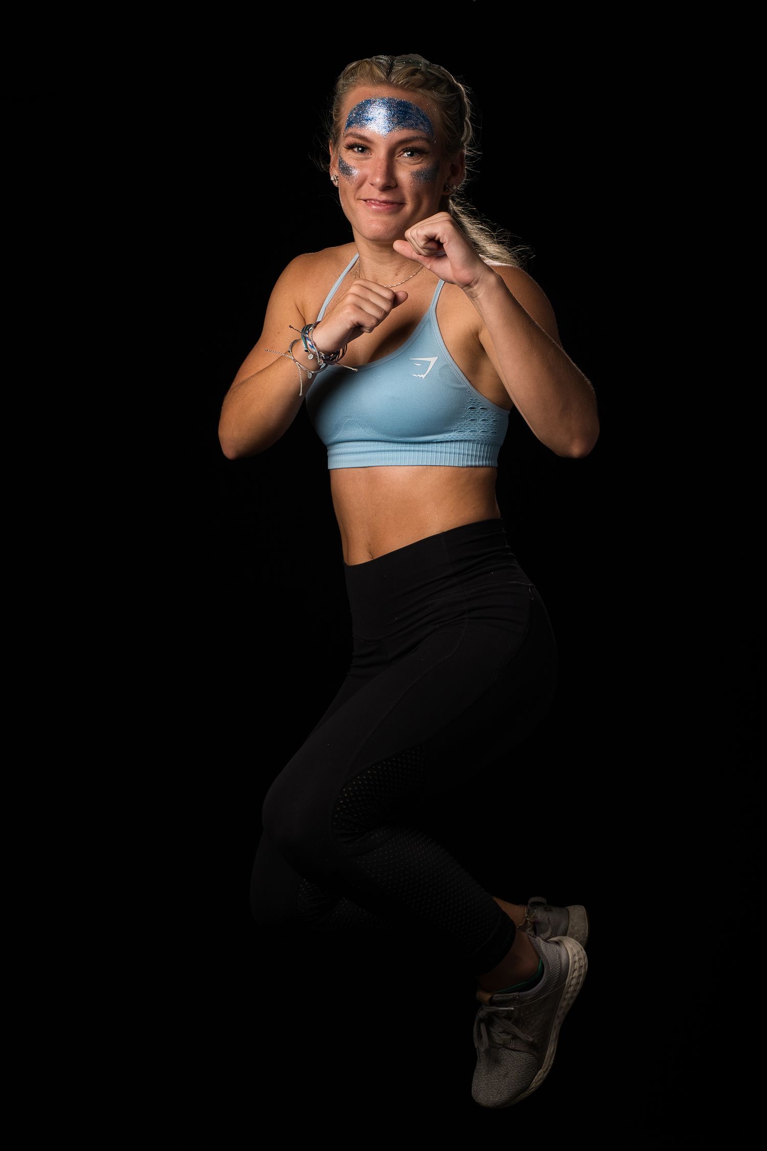 A fit blonde woman in a blue sports bra and black leggings poses in a fighting stance on black background.